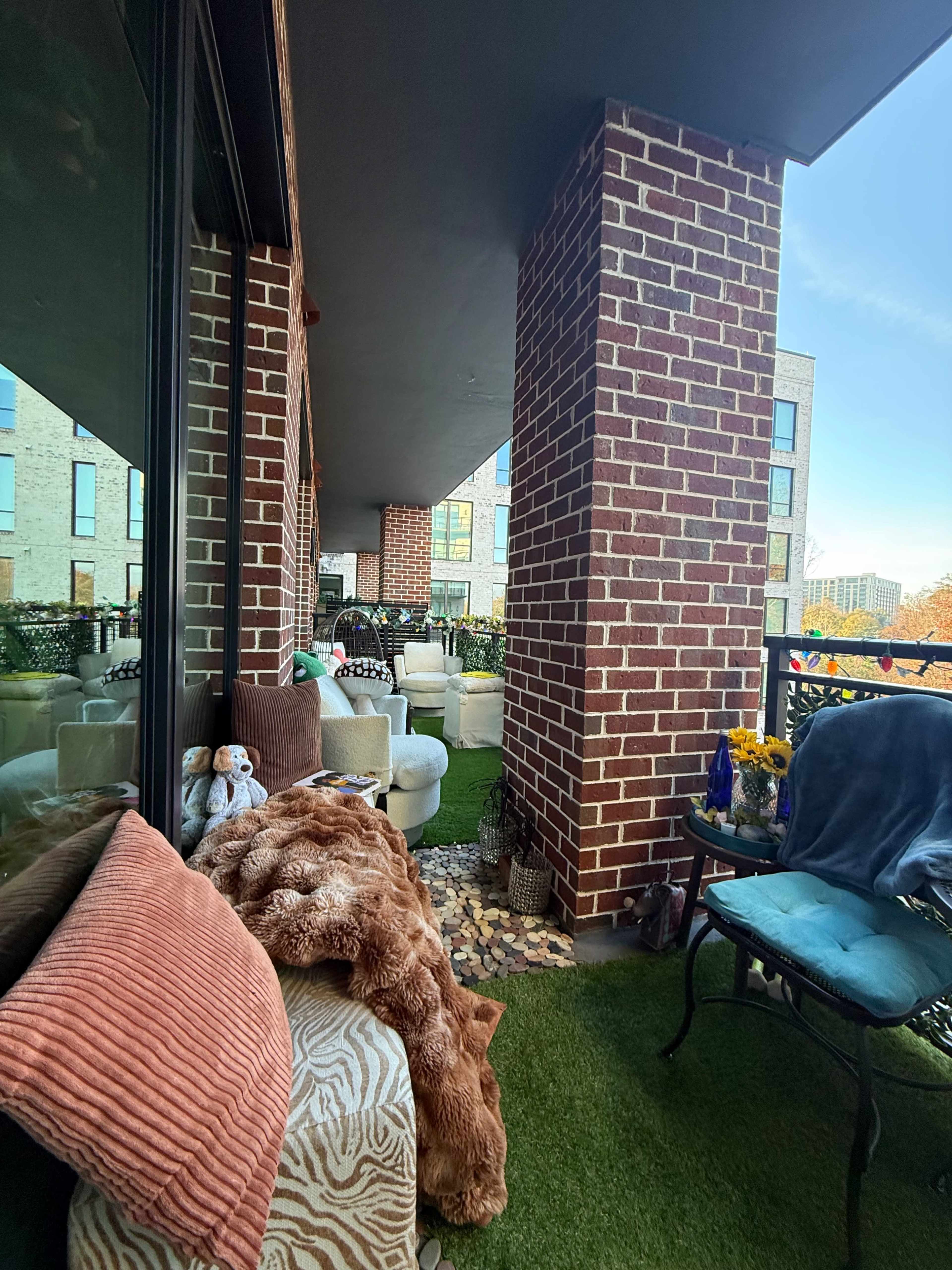 The image shows a cozy balcony area featuring a mix of plush seating, decorative pillows, and potted plants, with a view of a nearby building.