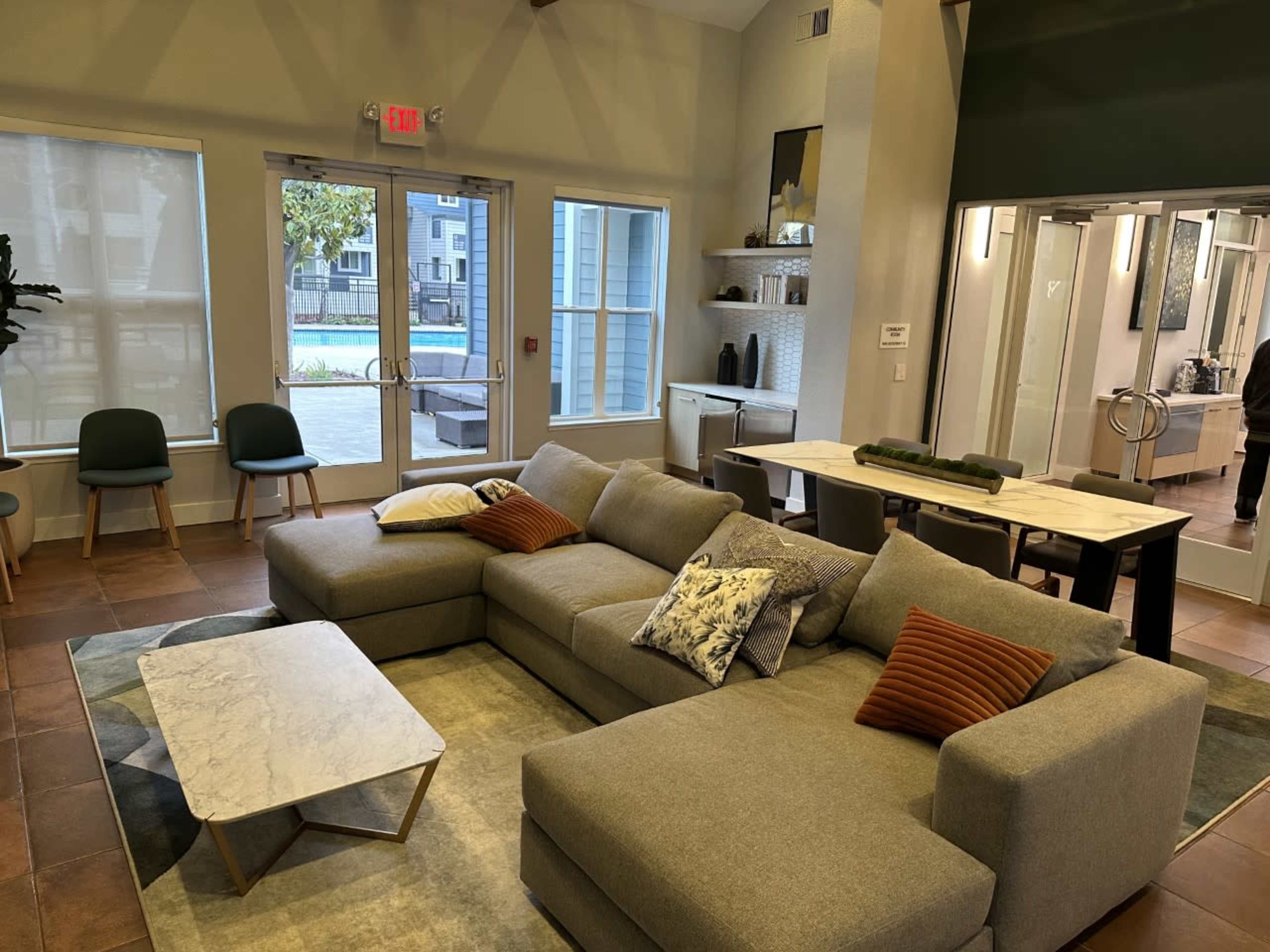 A modern lounge area with a large sectional sofa, a coffee table, and a dining area, featuring large windows that overlook an outdoor pool.