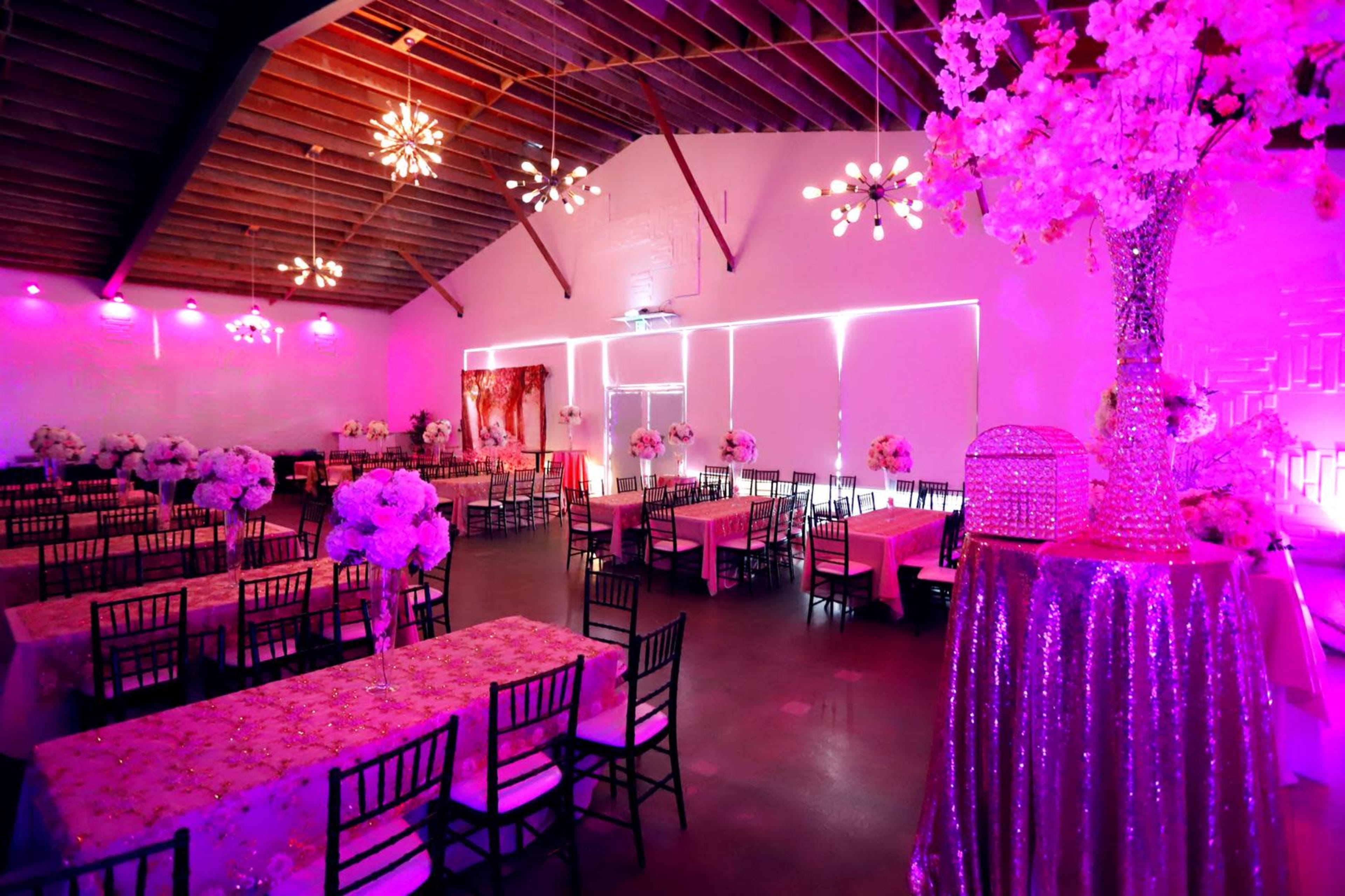 A banquet hall features long tables adorned with floral centerpieces, under soft pink lighting and decorative fixtures.