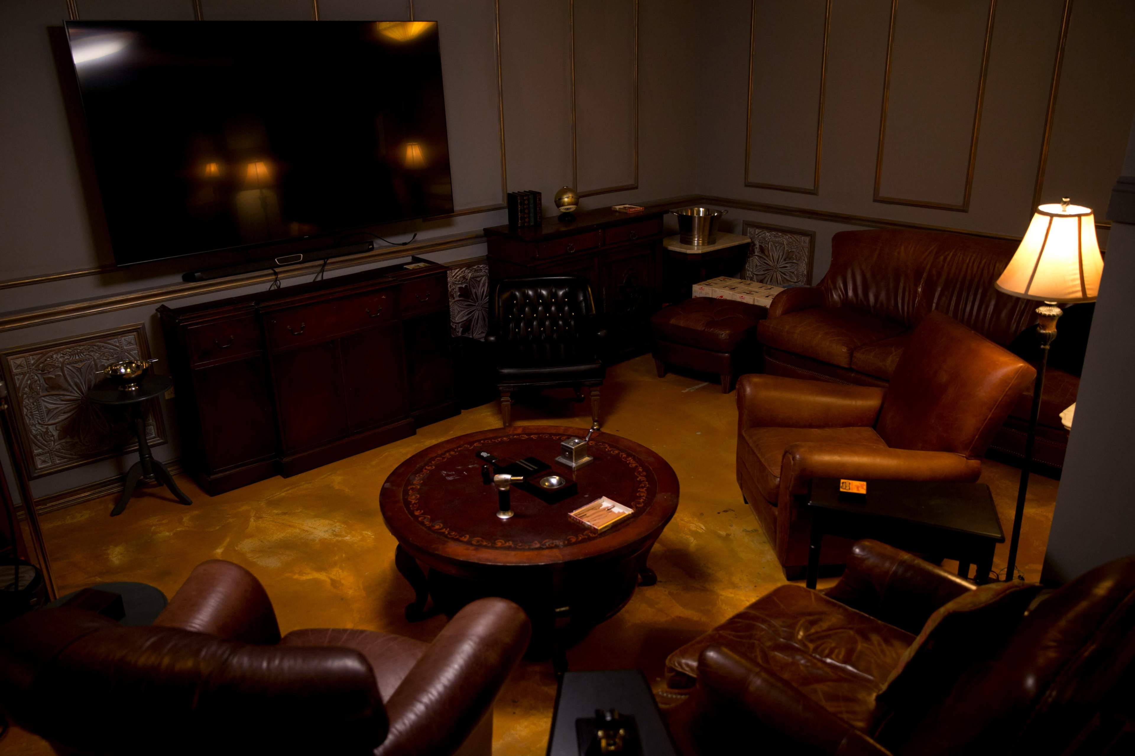 A dimly lit room features a large television mounted on the wall, surrounded by leather armchairs and a round coffee table.