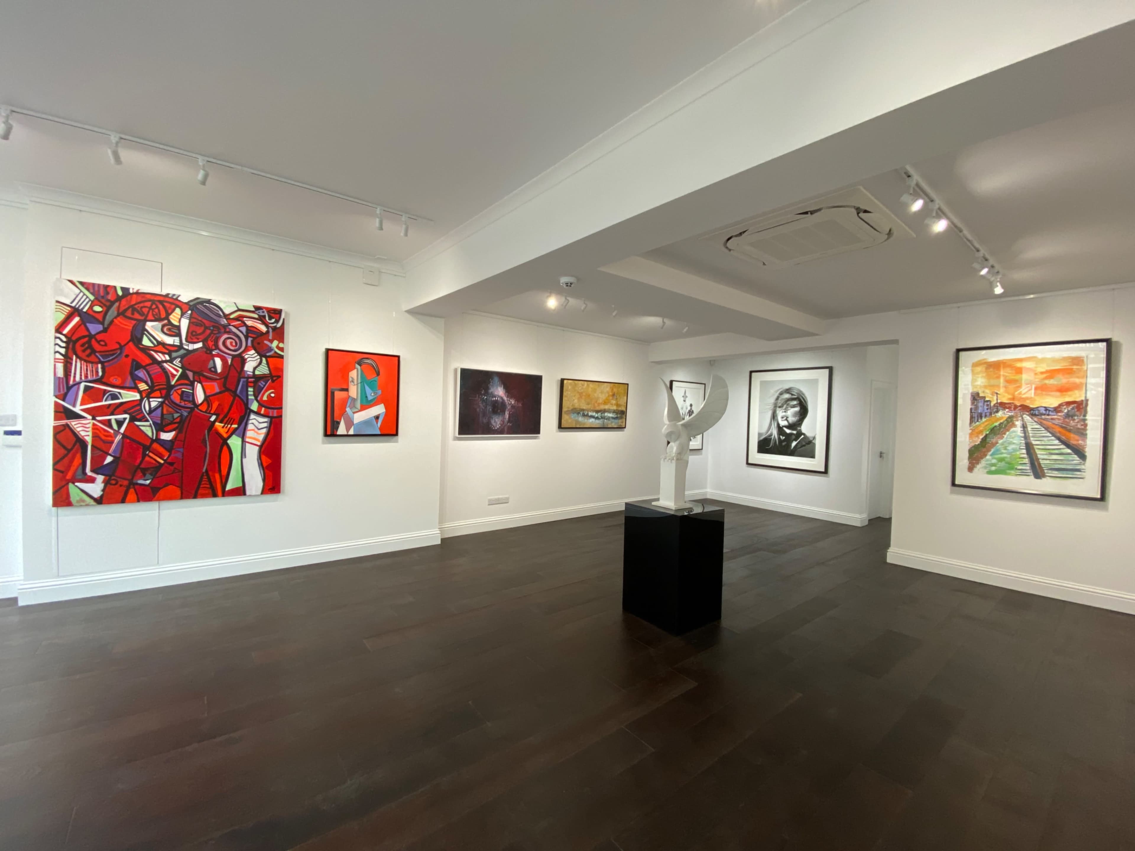 The image shows an art gallery with various paintings and a sculpture displayed on white walls and dark wooden flooring.