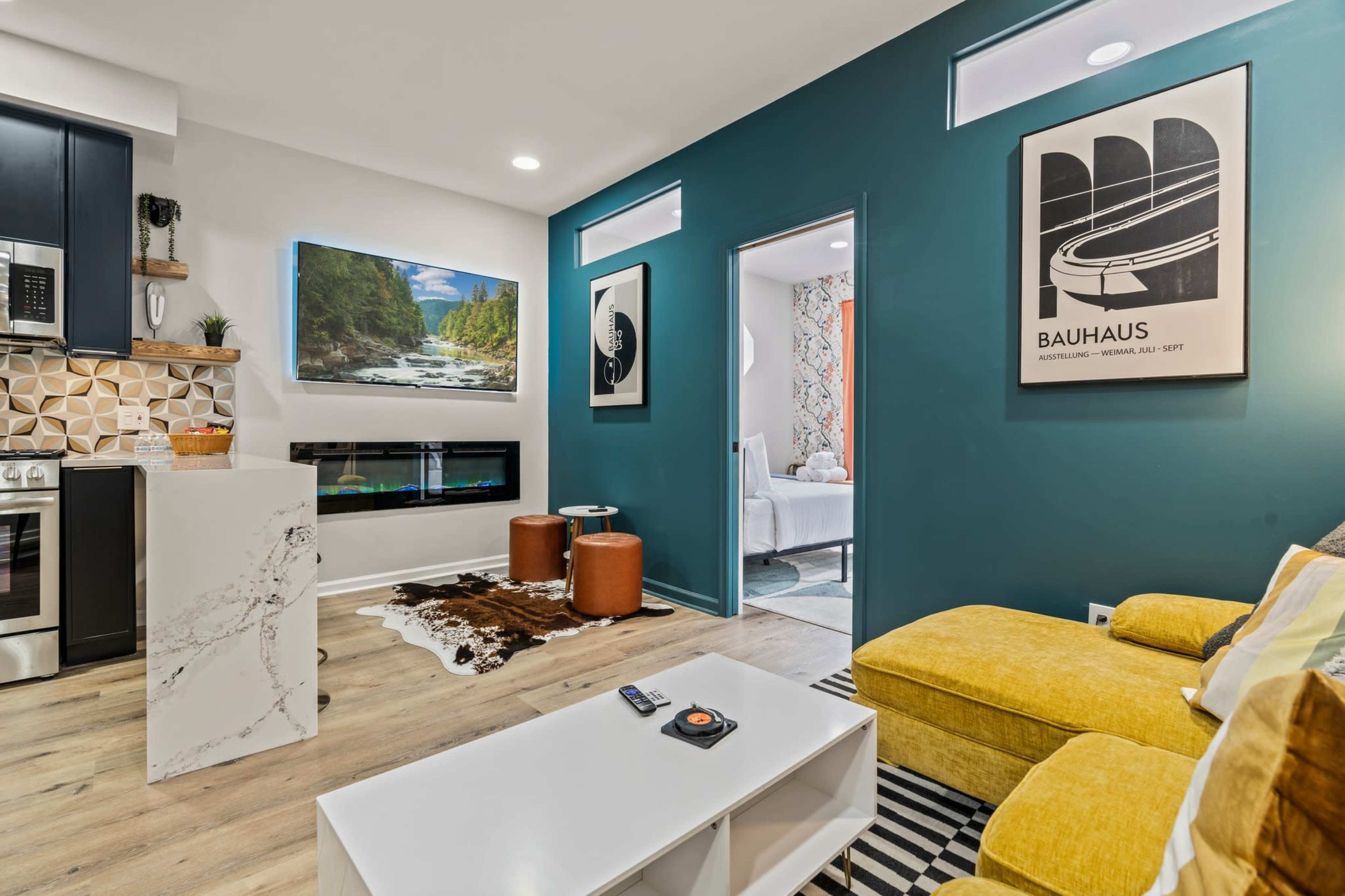 The image depicts a modern living room featuring a yellow couch, a white coffee table, and a wall-mounted television, with an open layout connecting to other areas.
