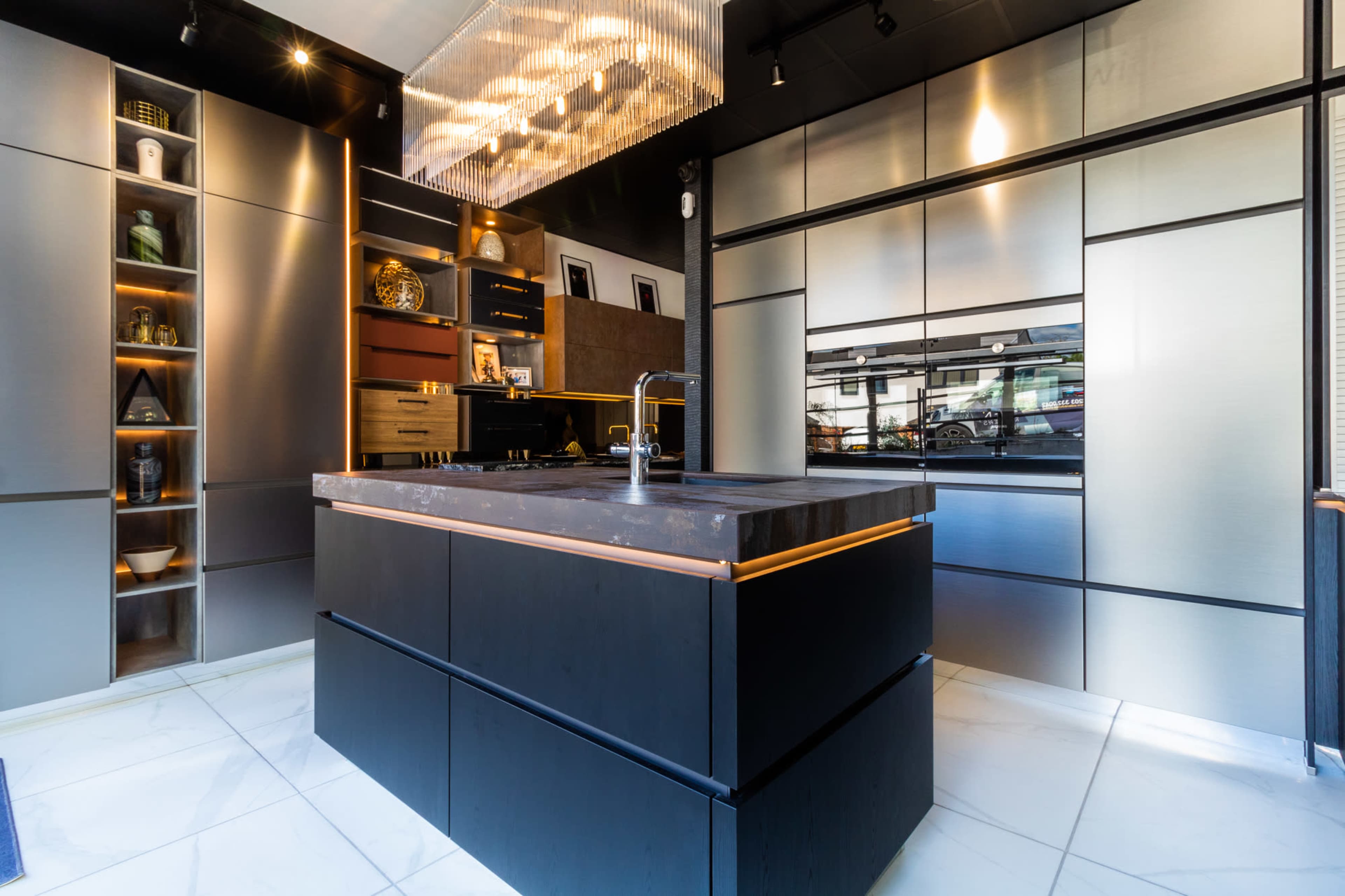 A modern kitchen features a central island with a dark countertop, sleek cabinetry, and a row of pendant lights above.