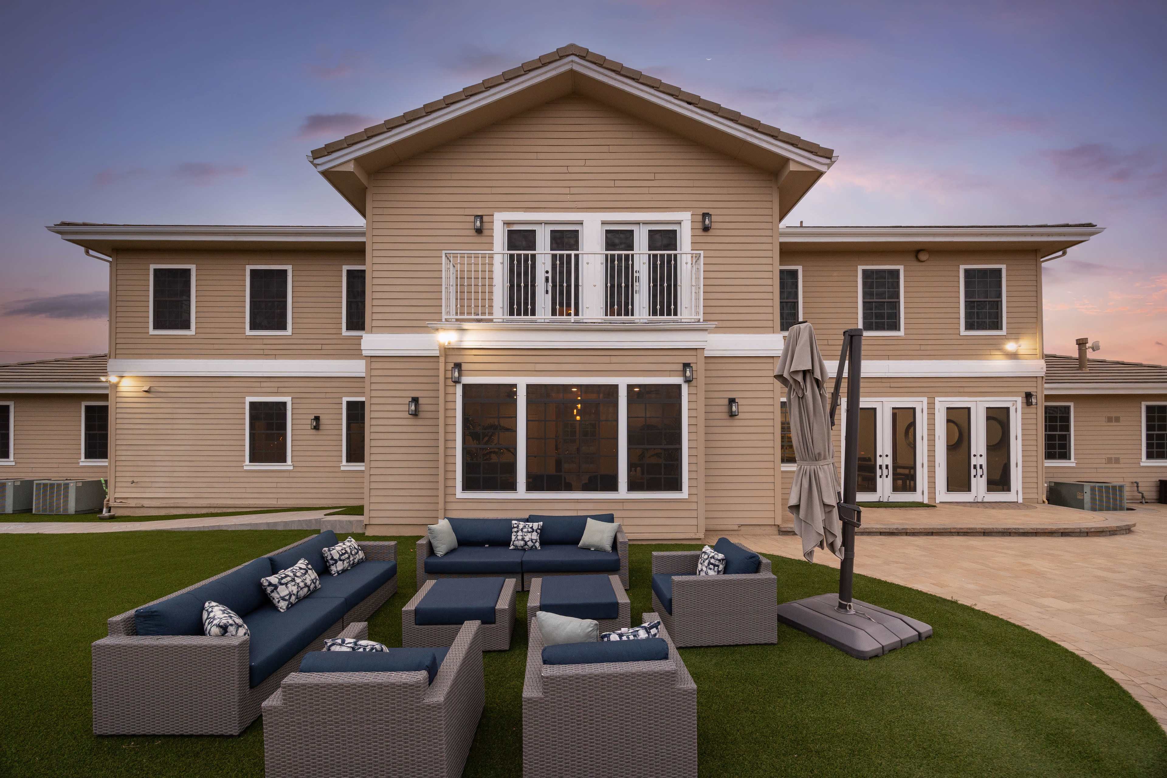 The image shows a large two-story wooden house with a spacious backyard featuring lounge seating arranged in a circular formation.