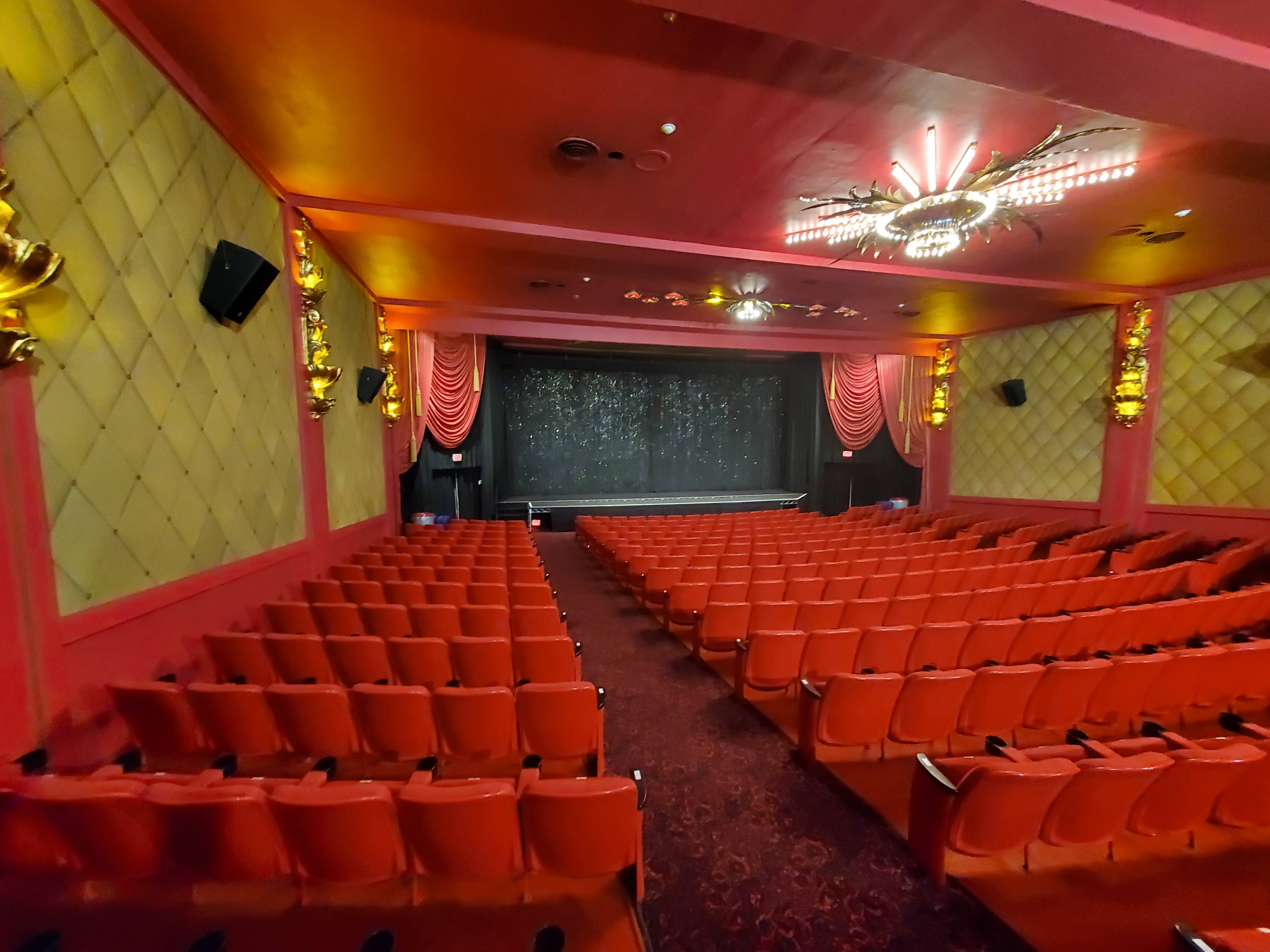 The image features an empty theater with red seats facing a stage draped in dark fabric, illuminated by ornate lighting above.