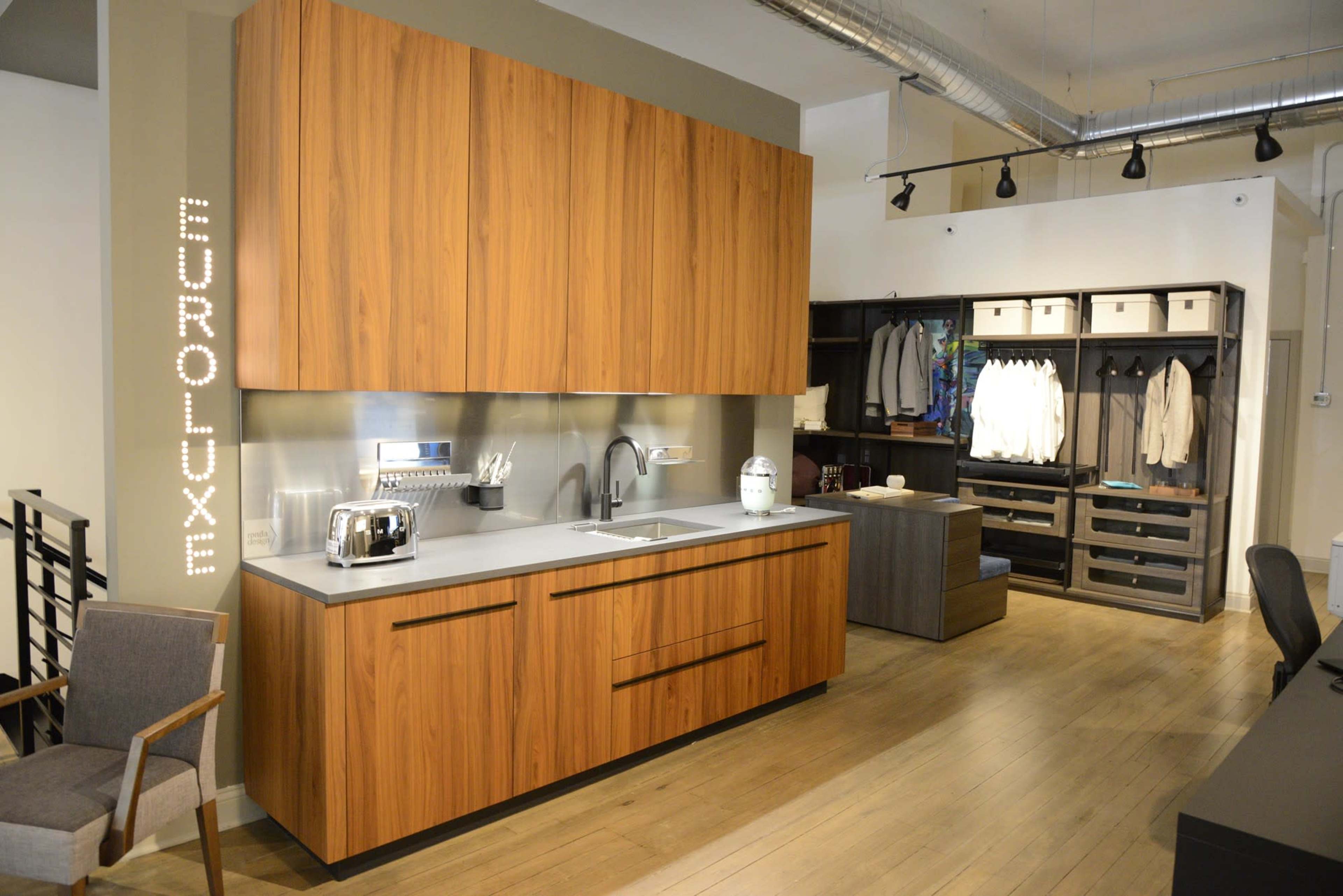 The image shows a modern kitchen display with wooden cabinetry, a stainless steel sink, and a countertop, alongside a clothing storage area featuring hanging garments.