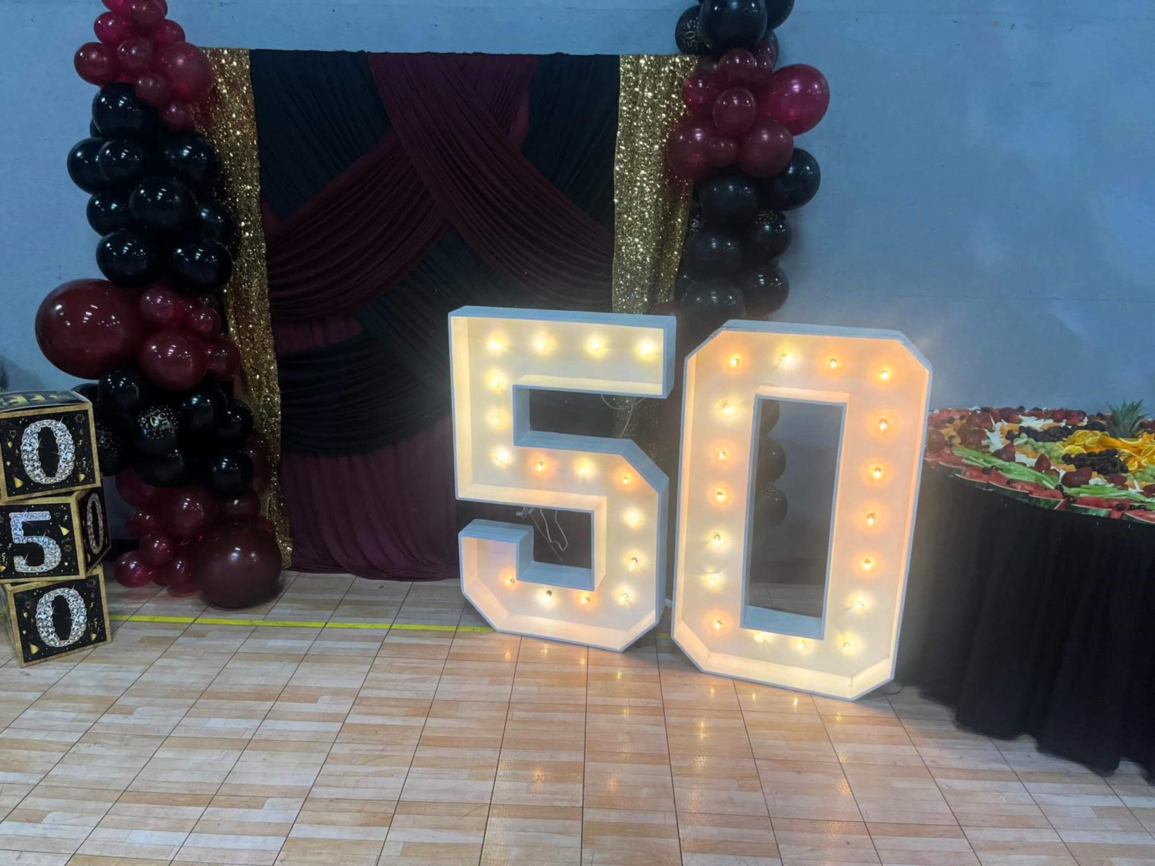 The image shows a decorated event space featuring large illuminated numbers "50," a backdrop with draped fabric and balloons, and a table with a floral arrangement.