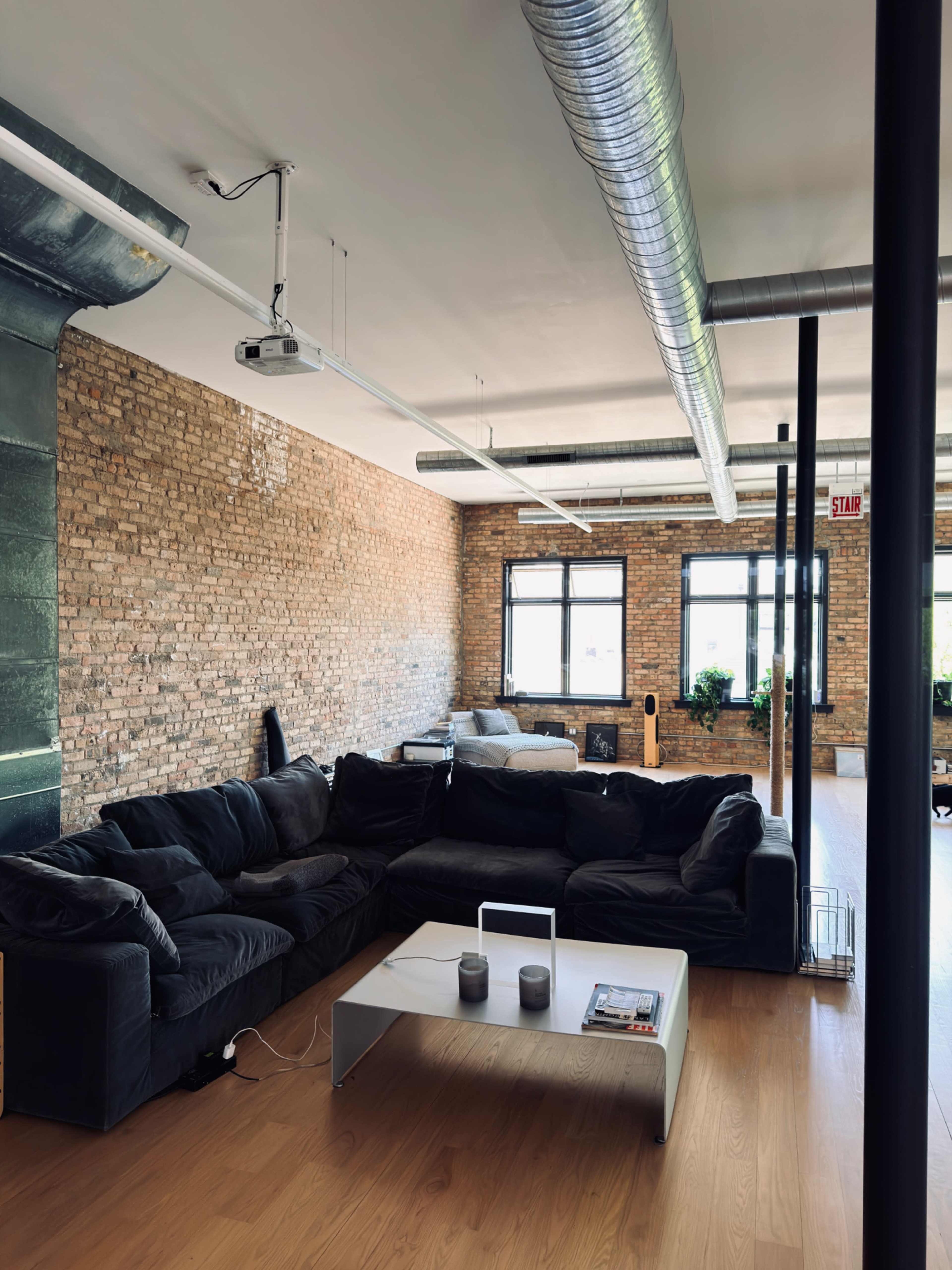 The image shows a spacious living area with a black sectional sofa, a minimalist coffee table, and exposed brick walls.