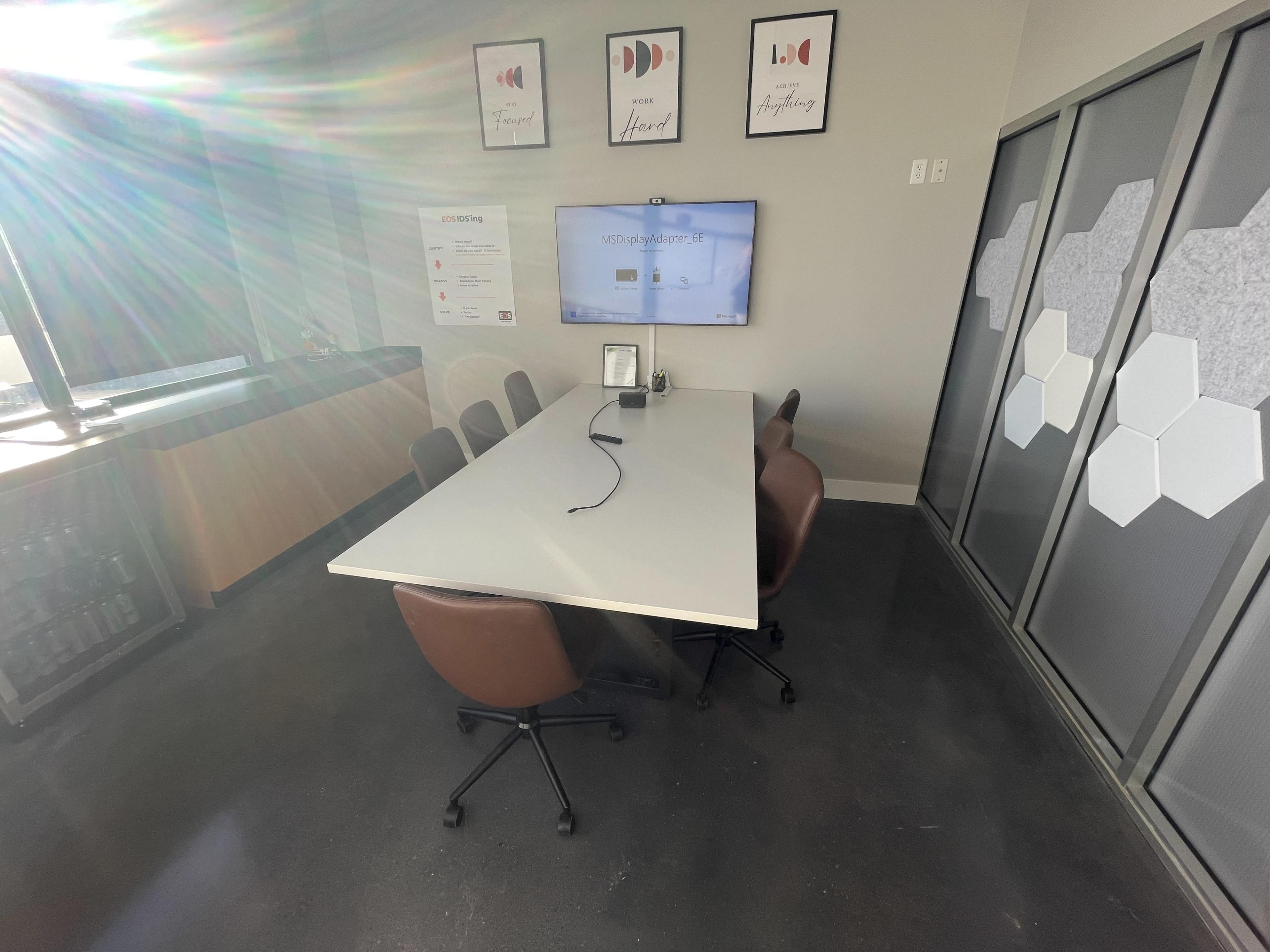The image shows a modern conference room with a rectangular table, four brown chairs, a wall-mounted screen, and several framed artworks.