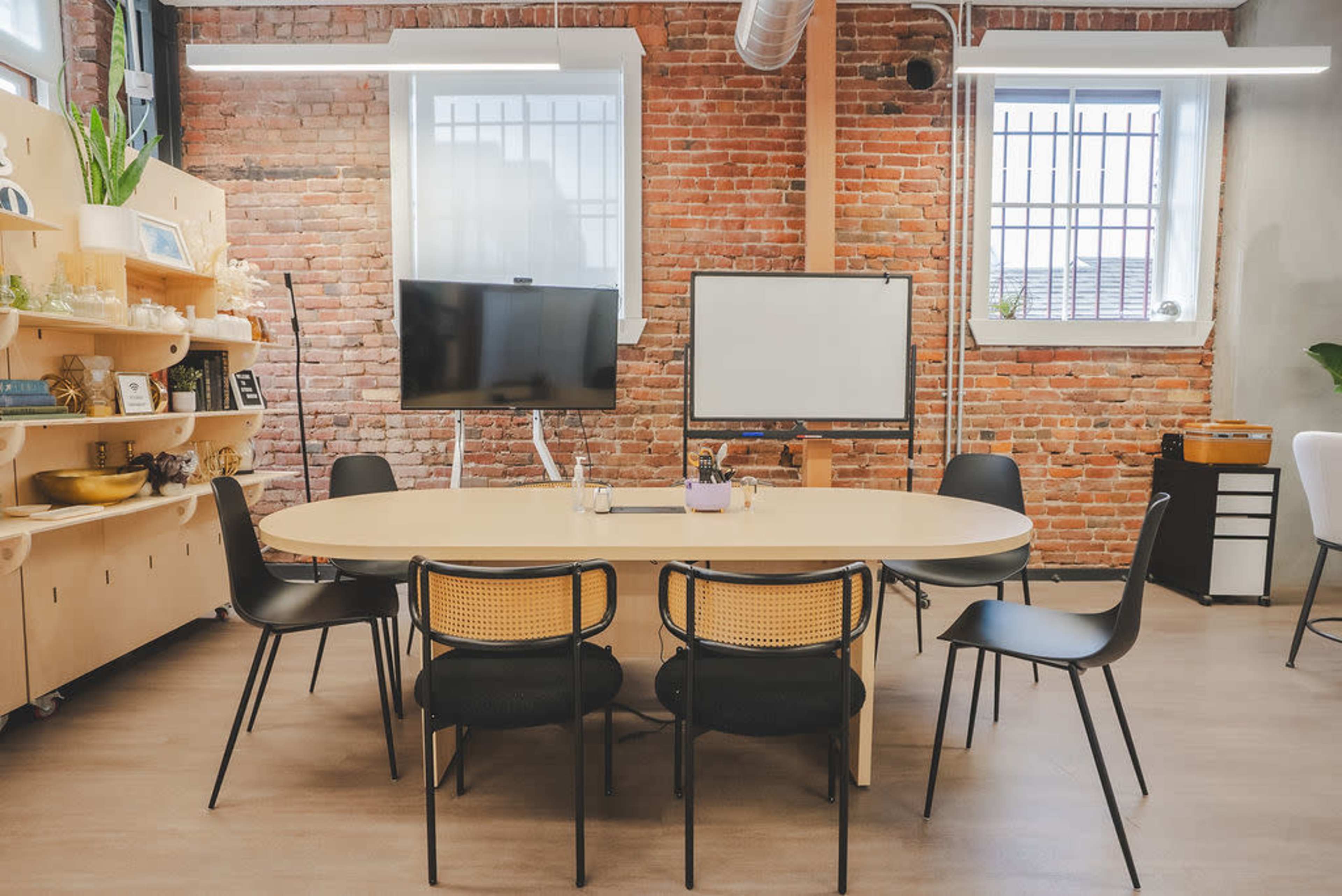A modern conference room features a large oval table surrounded by black chairs, with a television and whiteboard visible on the brick wall.