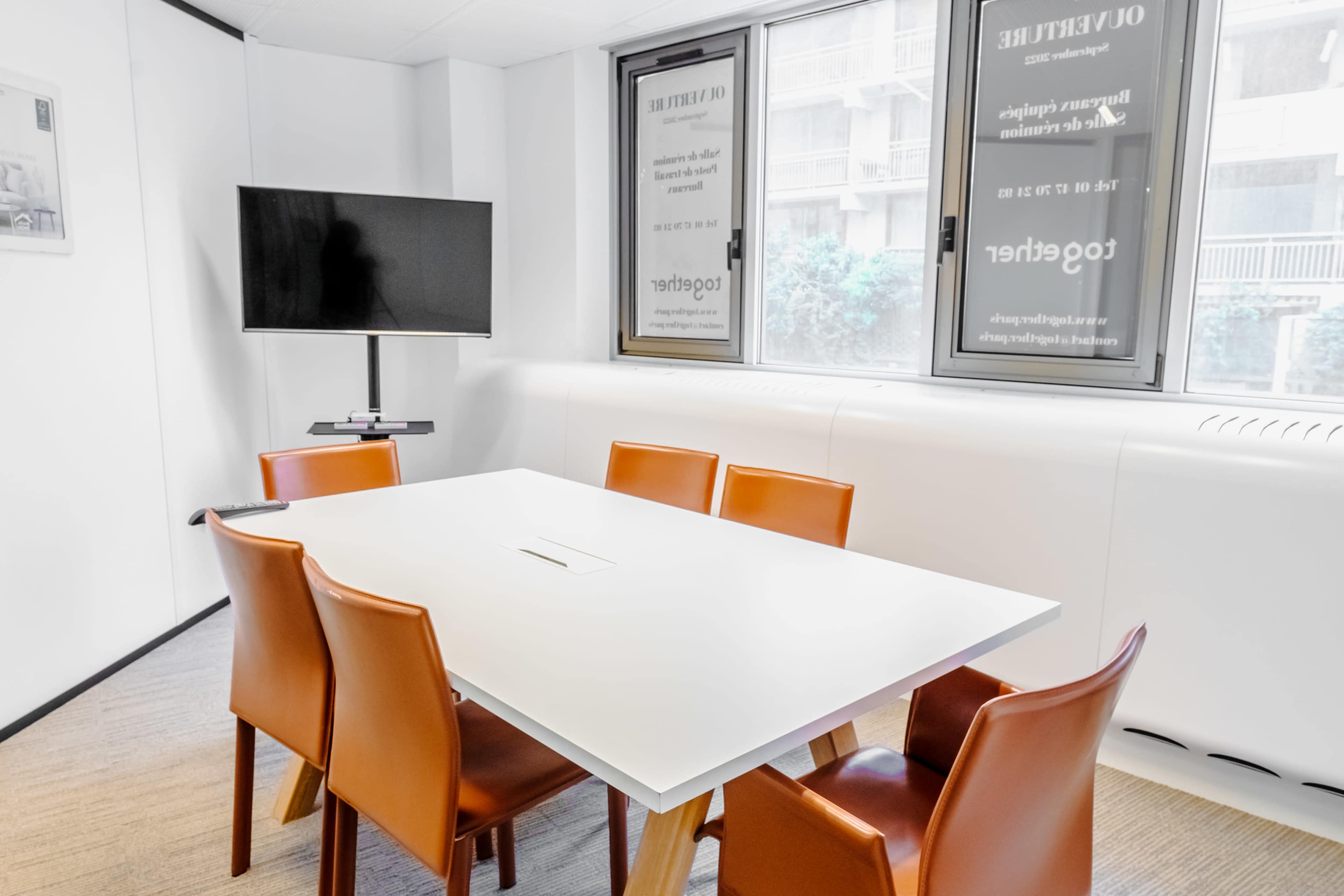 A modern conference room features a rectangular white table surrounded by six orange chairs, with a television mounted on the wall and large windows offering natural light.