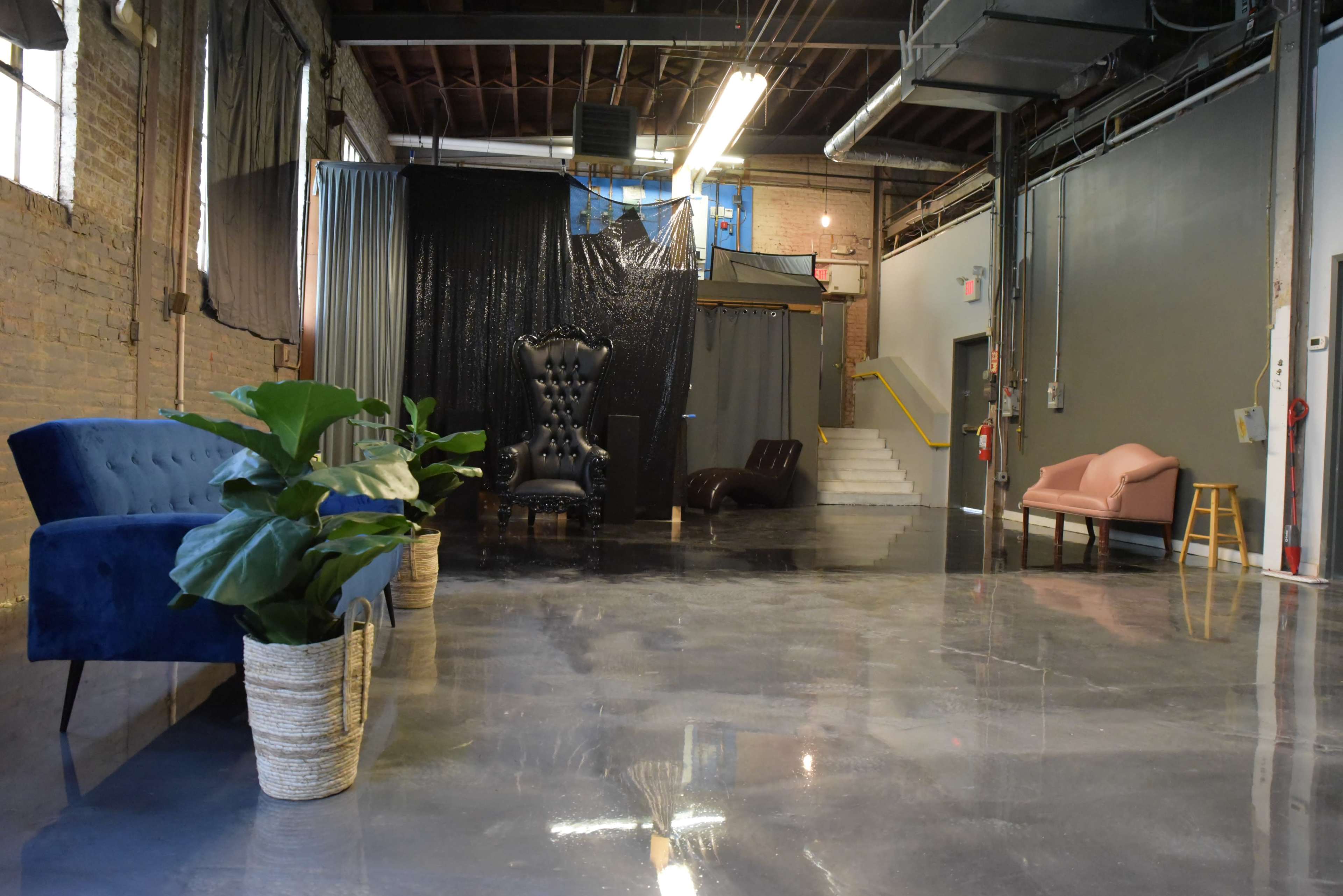 A spacious industrial-style room featuring various seating arrangements, including a blue couch, a pink sofa, and a black chair, with polished concrete floors and exposed walls.