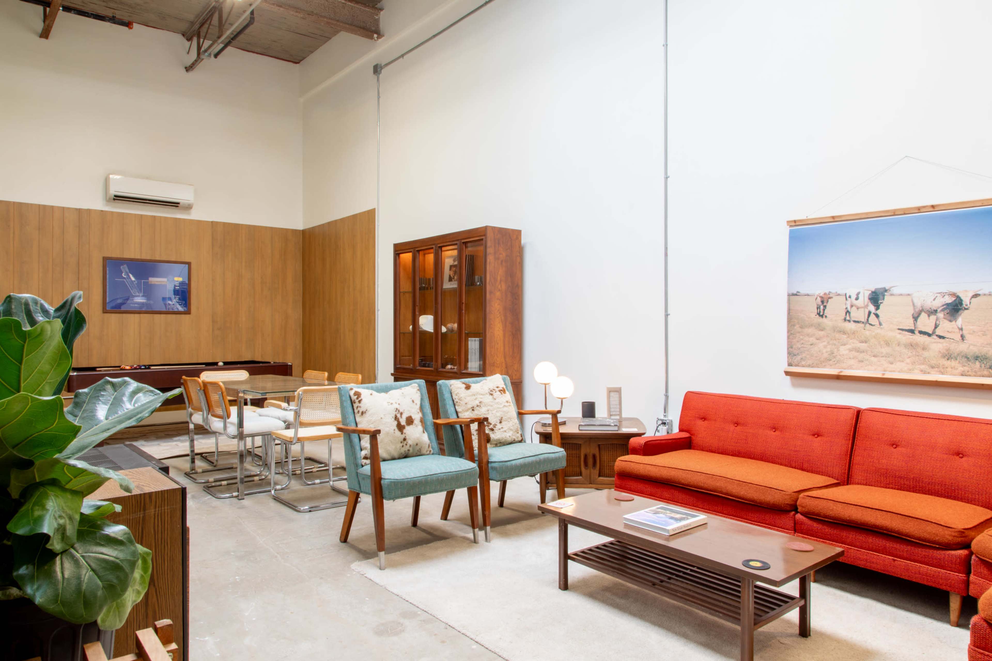 A modern, minimalist interior featuring a red sofa, two patterned armchairs, a wooden dining table, and a large wall-mounted photograph.