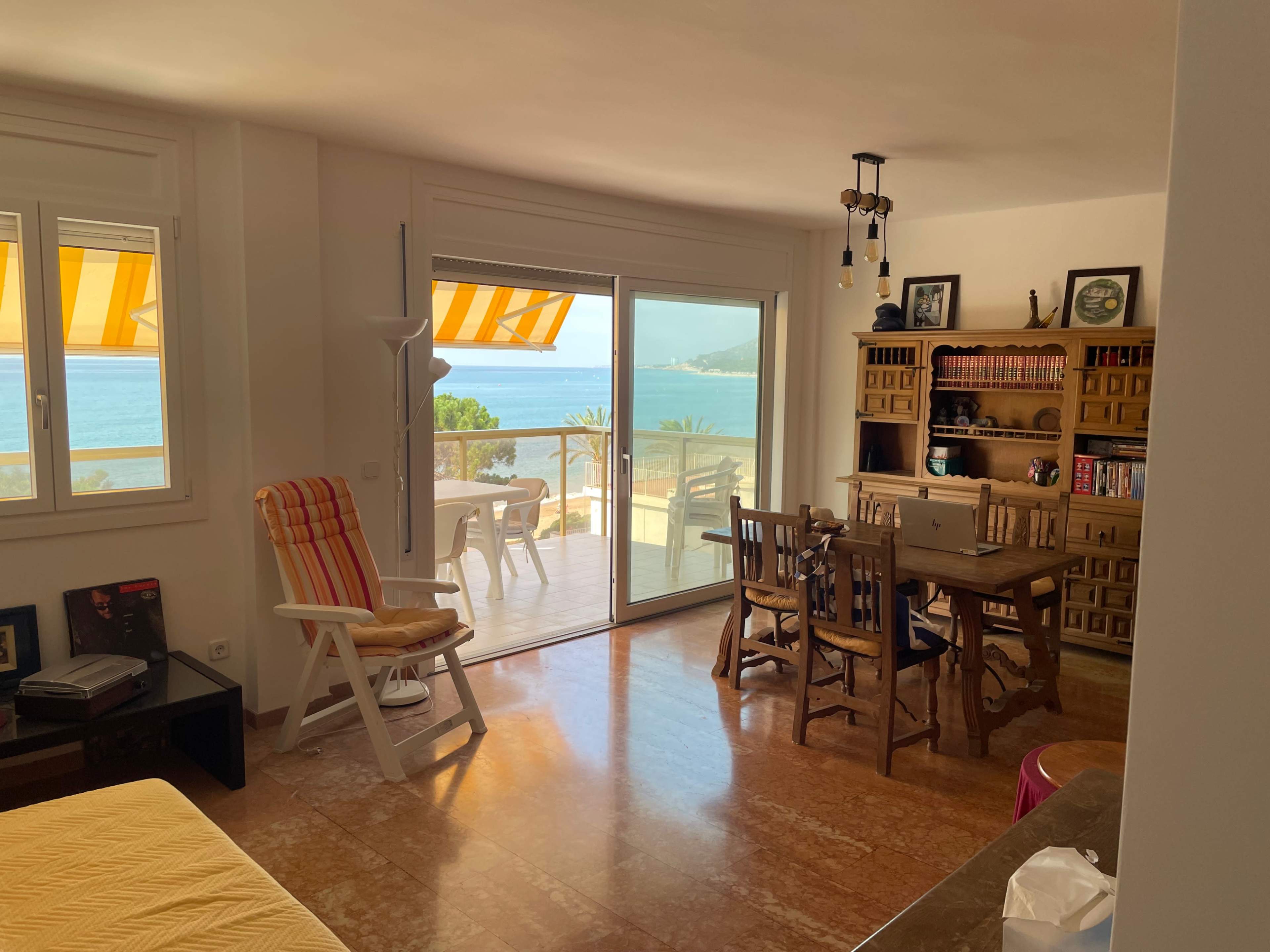 A brightly lit room features a seating area with a wooden table and chairs, and a large window overlooks a coastal view.