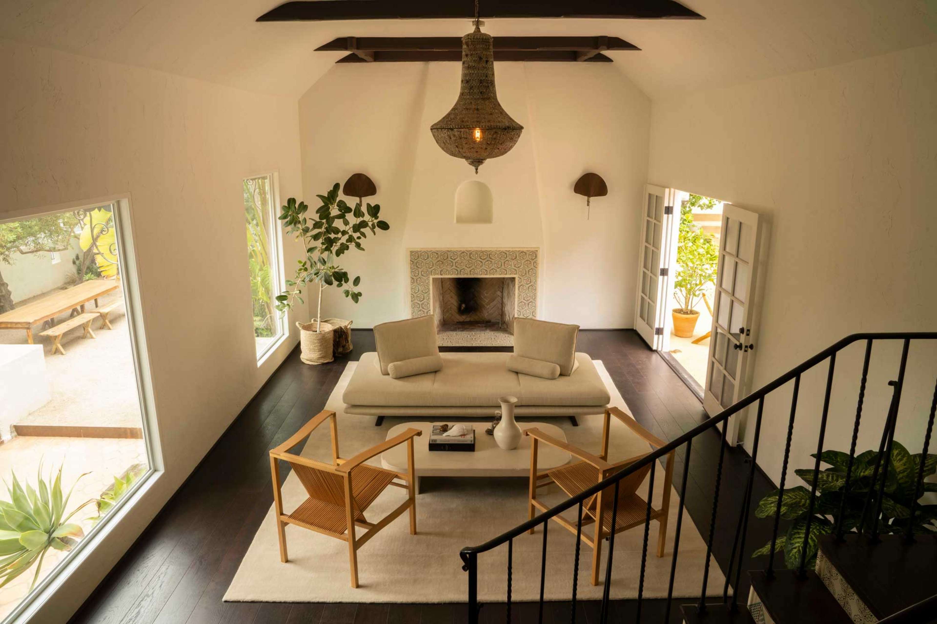 A bright, airy living room features a sofa, two wooden chairs, and a central table, with large windows and a fireplace.
