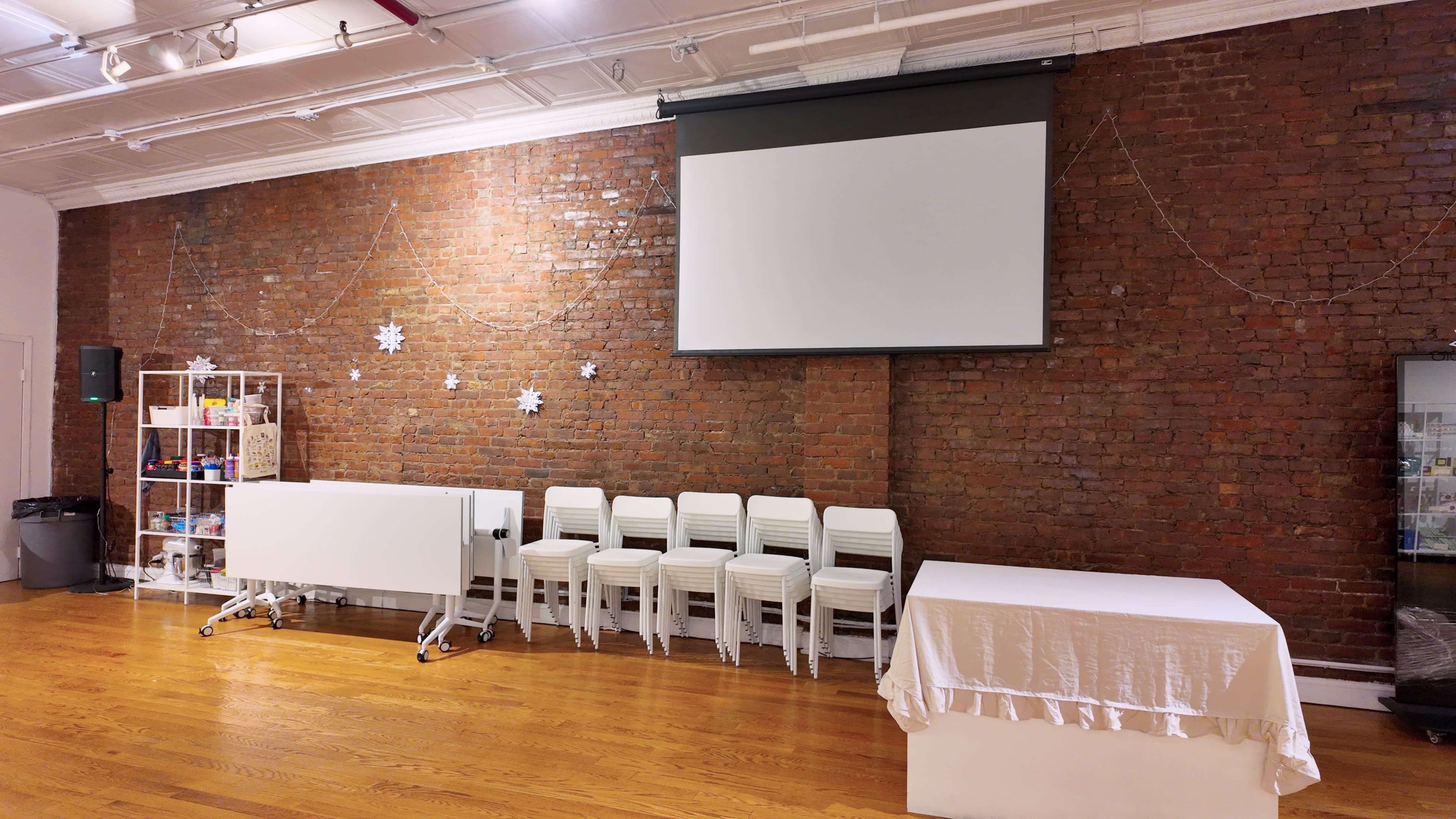 The image shows an interior space with a brick wall, a large blank projection screen, stacked white chairs, a table covered with a white cloth, and shelves with various items.