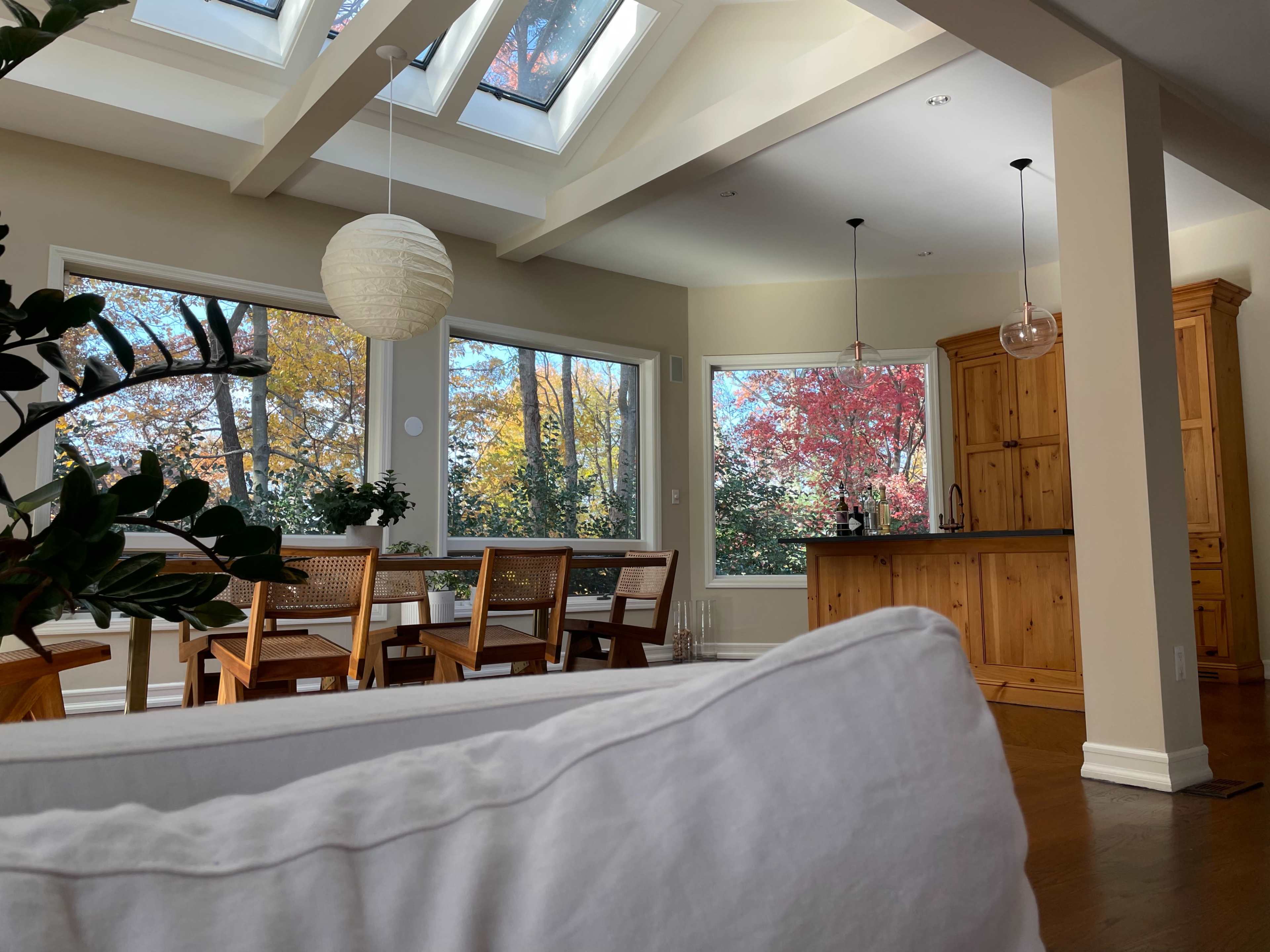 A bright interior space features a dining area with wooden chairs and a view of colorful autumn trees through large windows, accompanied by a kitchen outfitted with wooden cabinetry.