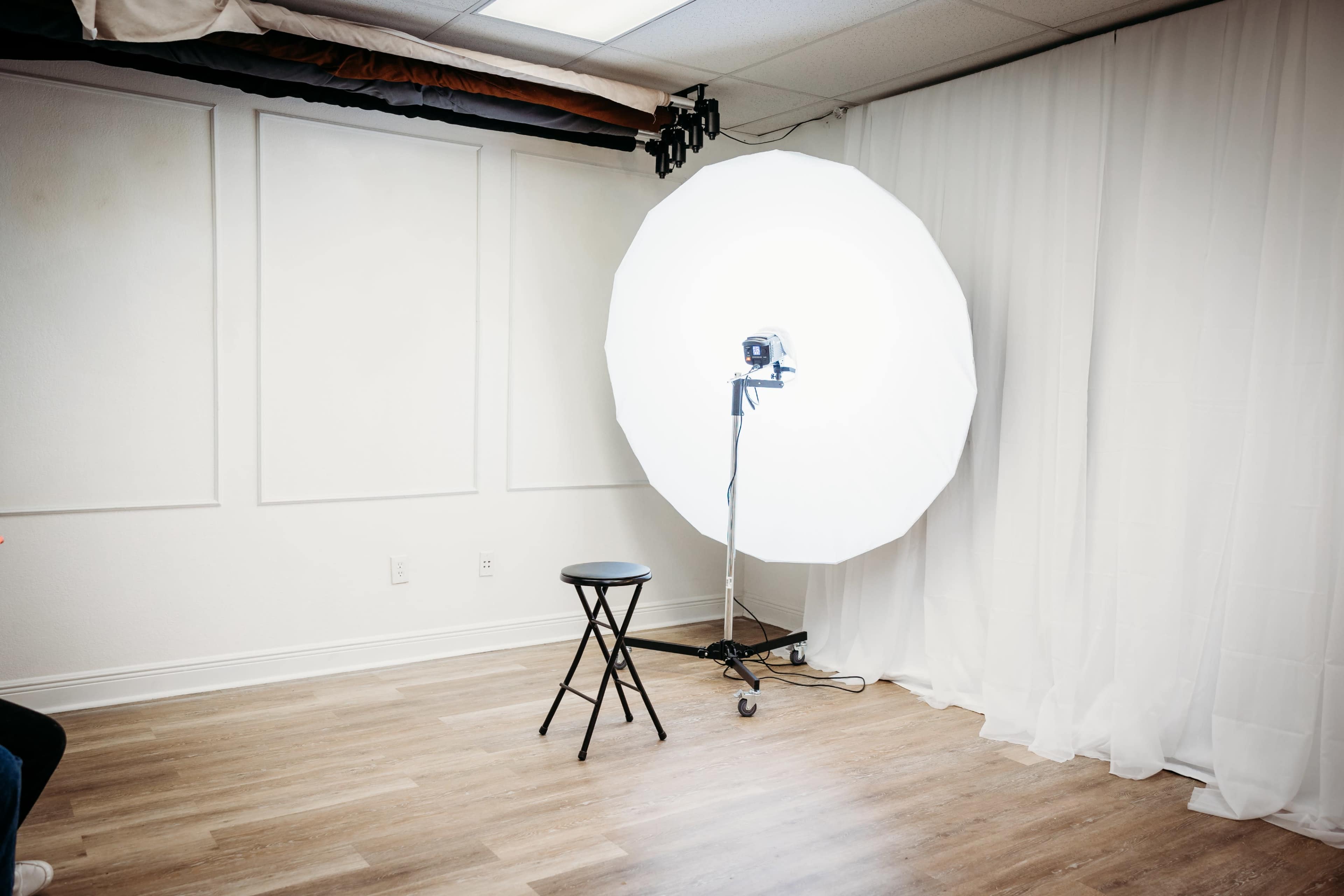 A photography setup featuring a large softbox light positioned near an empty black stool in a minimalistic studio space.