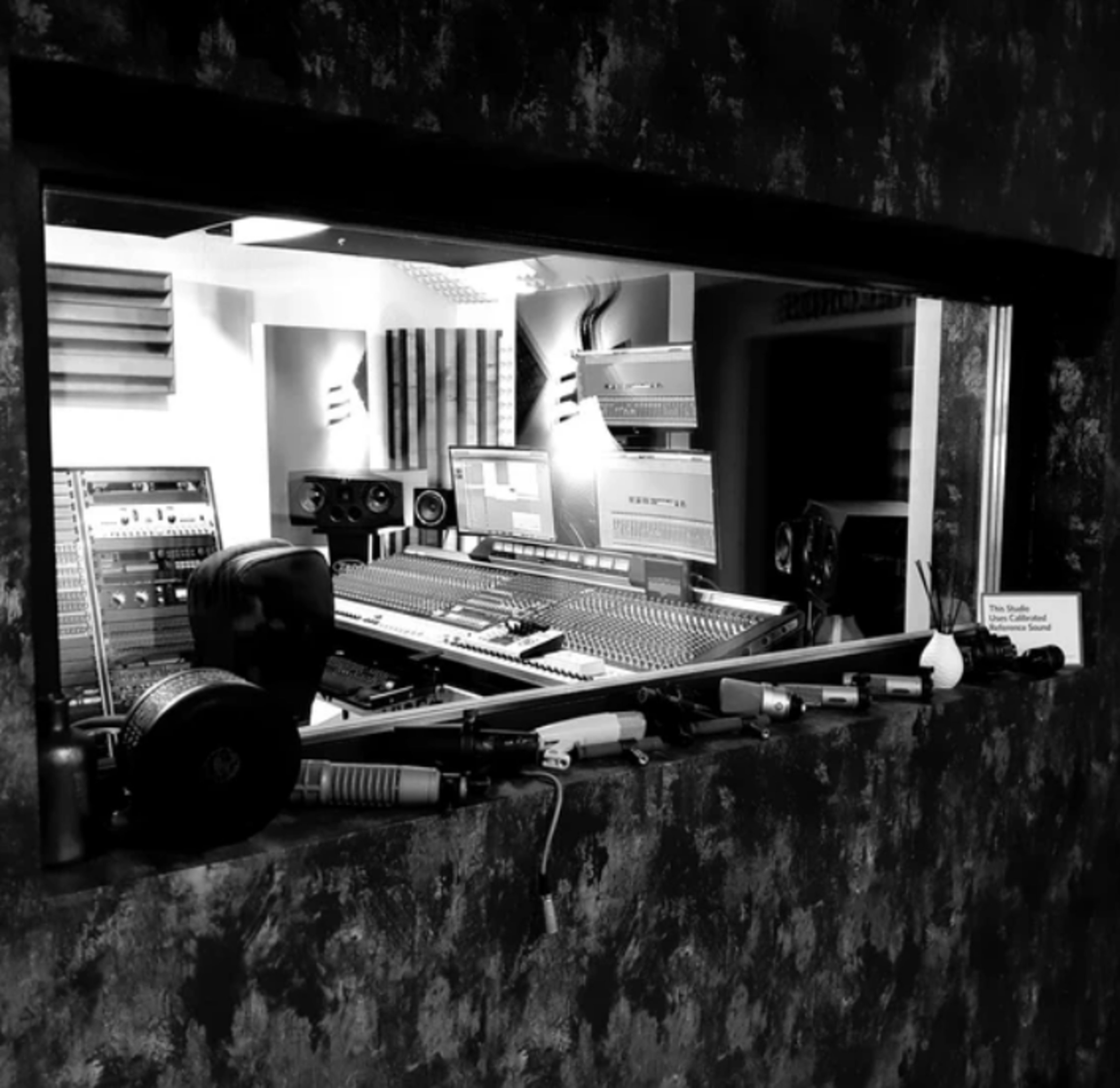 The image shows a sound recording studio featuring a mixing console, audio equipment, and various musical instruments visible through a window.