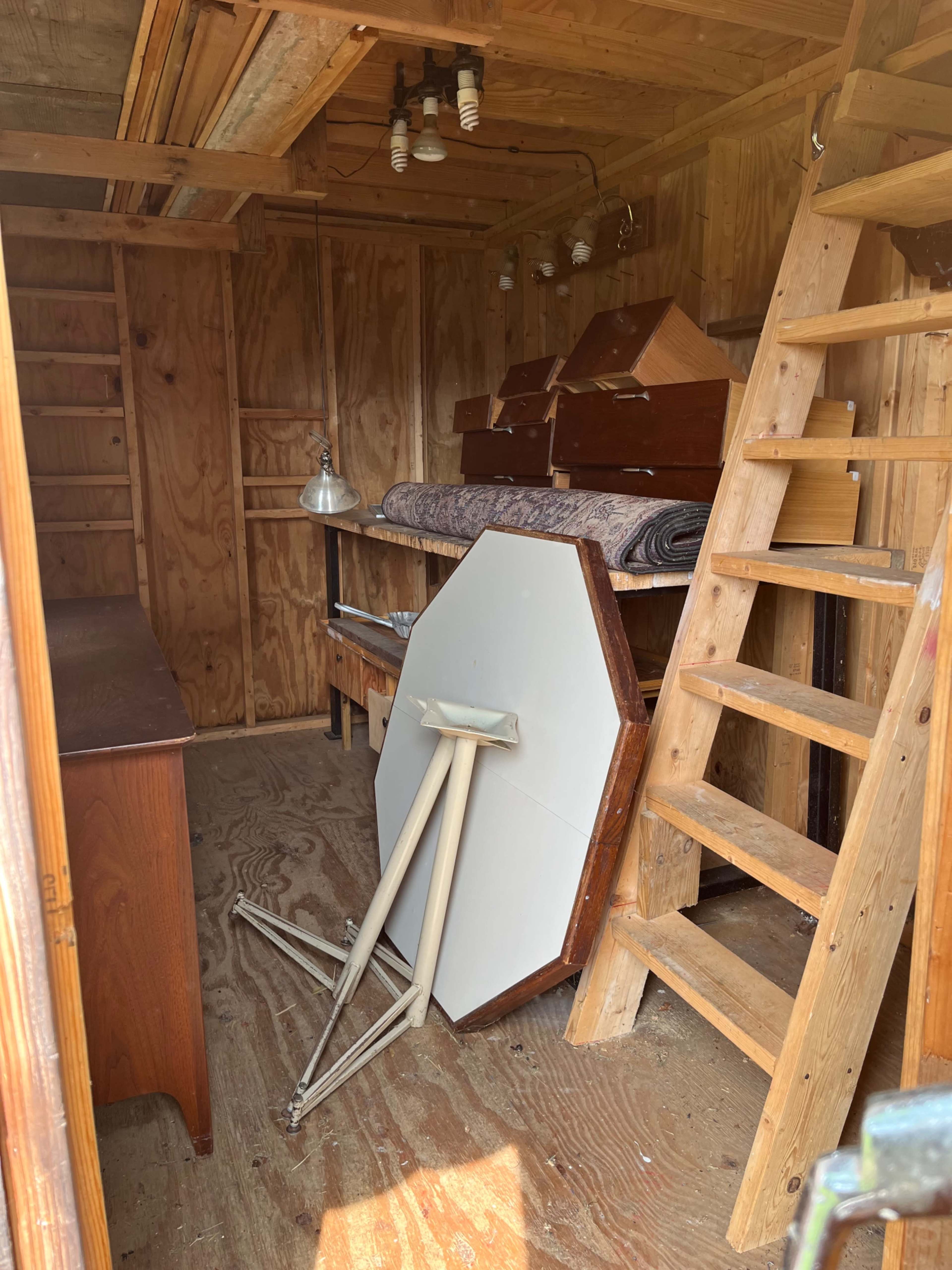 A wooden shed interior with stacked furniture, including a folding table, a lamp, and a wooden ladder against the wall.