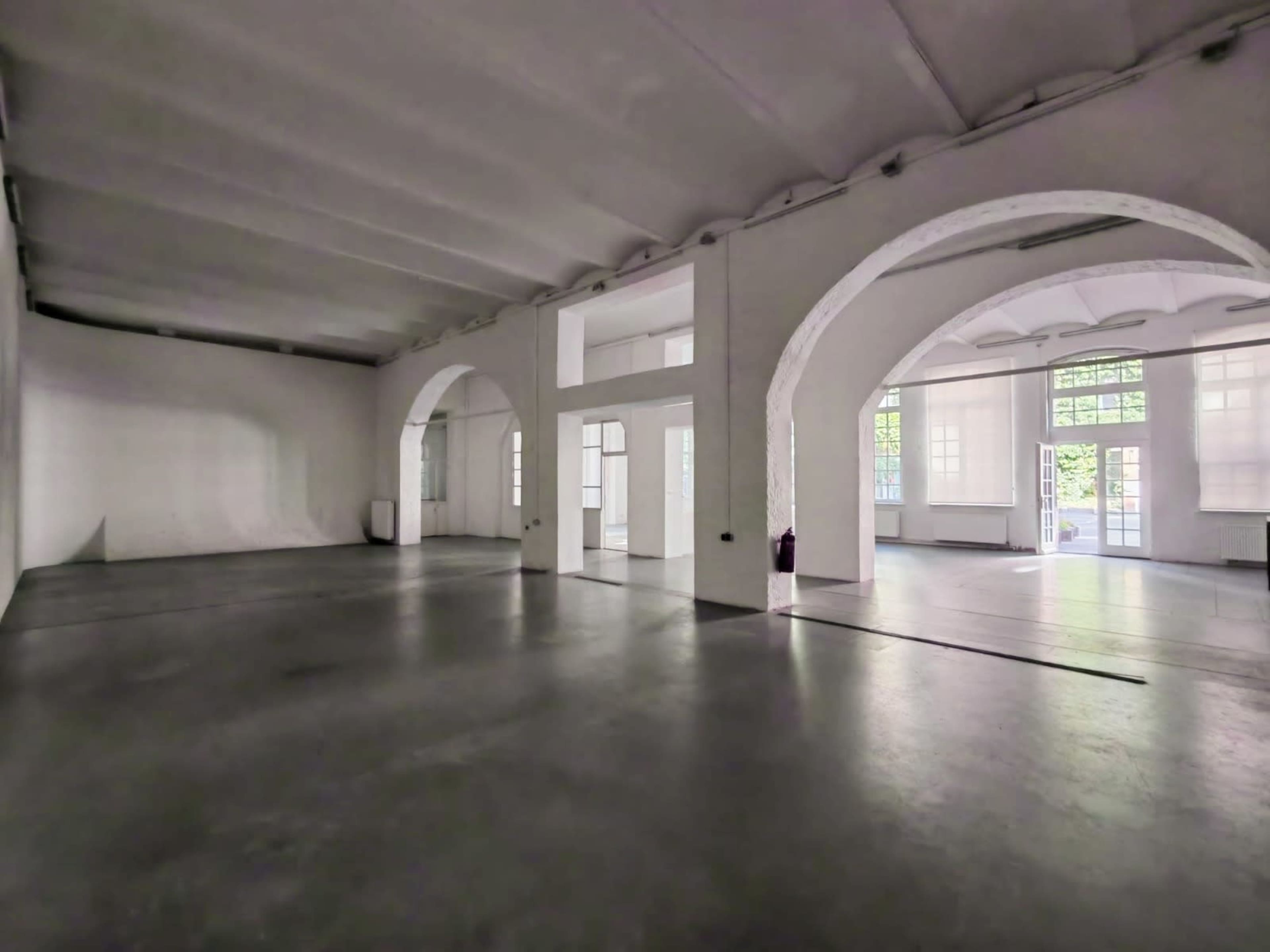 The image shows a spacious, empty room with high ceilings and large arched windows, featuring a simple concrete floor.