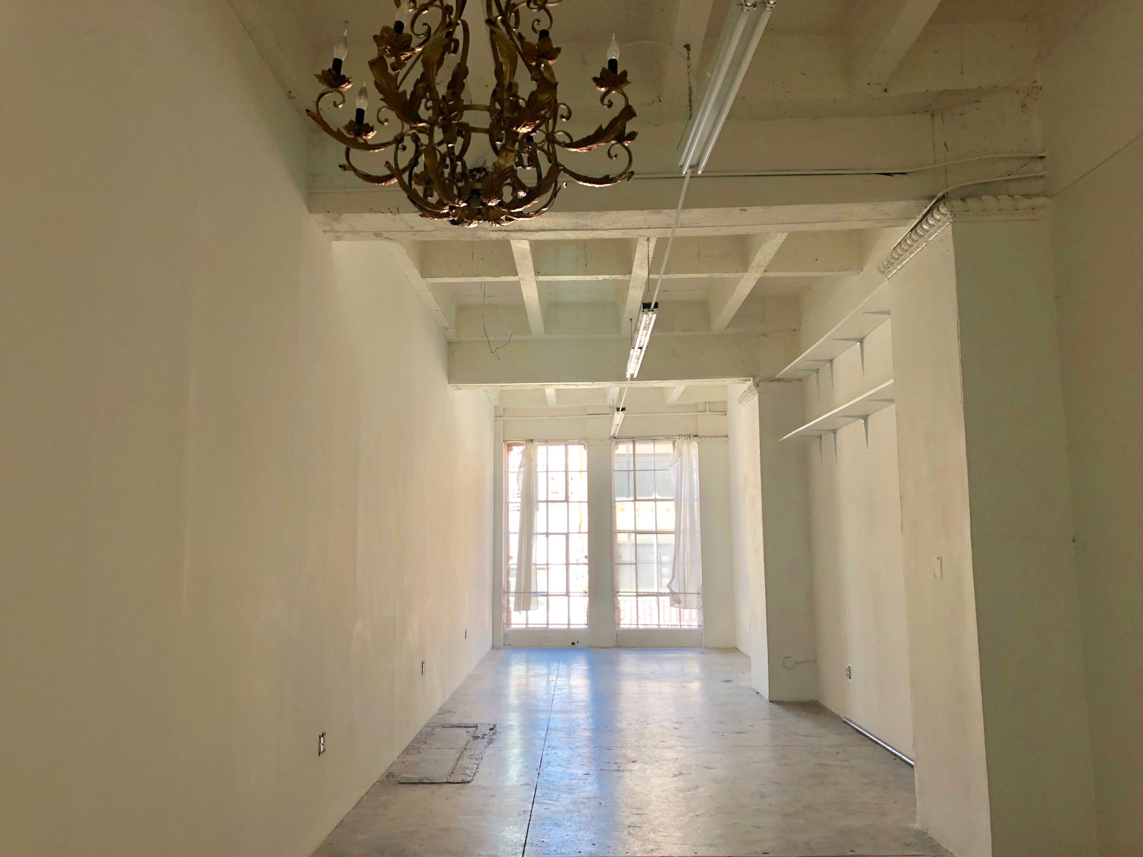 The image shows a spacious, empty room with high ceilings, a chandelier, and large windows allowing natural light to enter.
