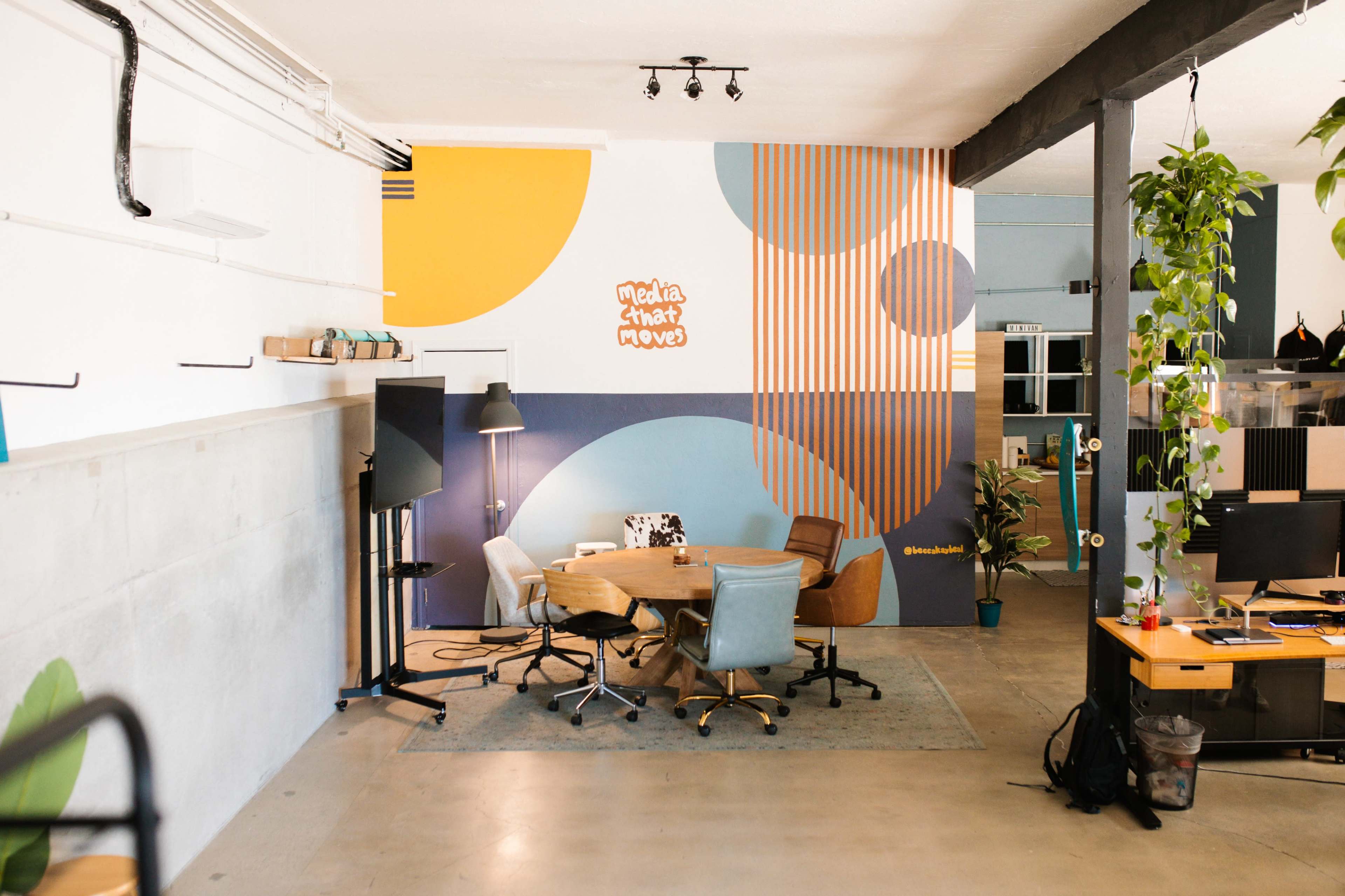 The image shows a modern office space with a circular table surrounded by chairs, colorful wall patterns, and a plant in the corner.