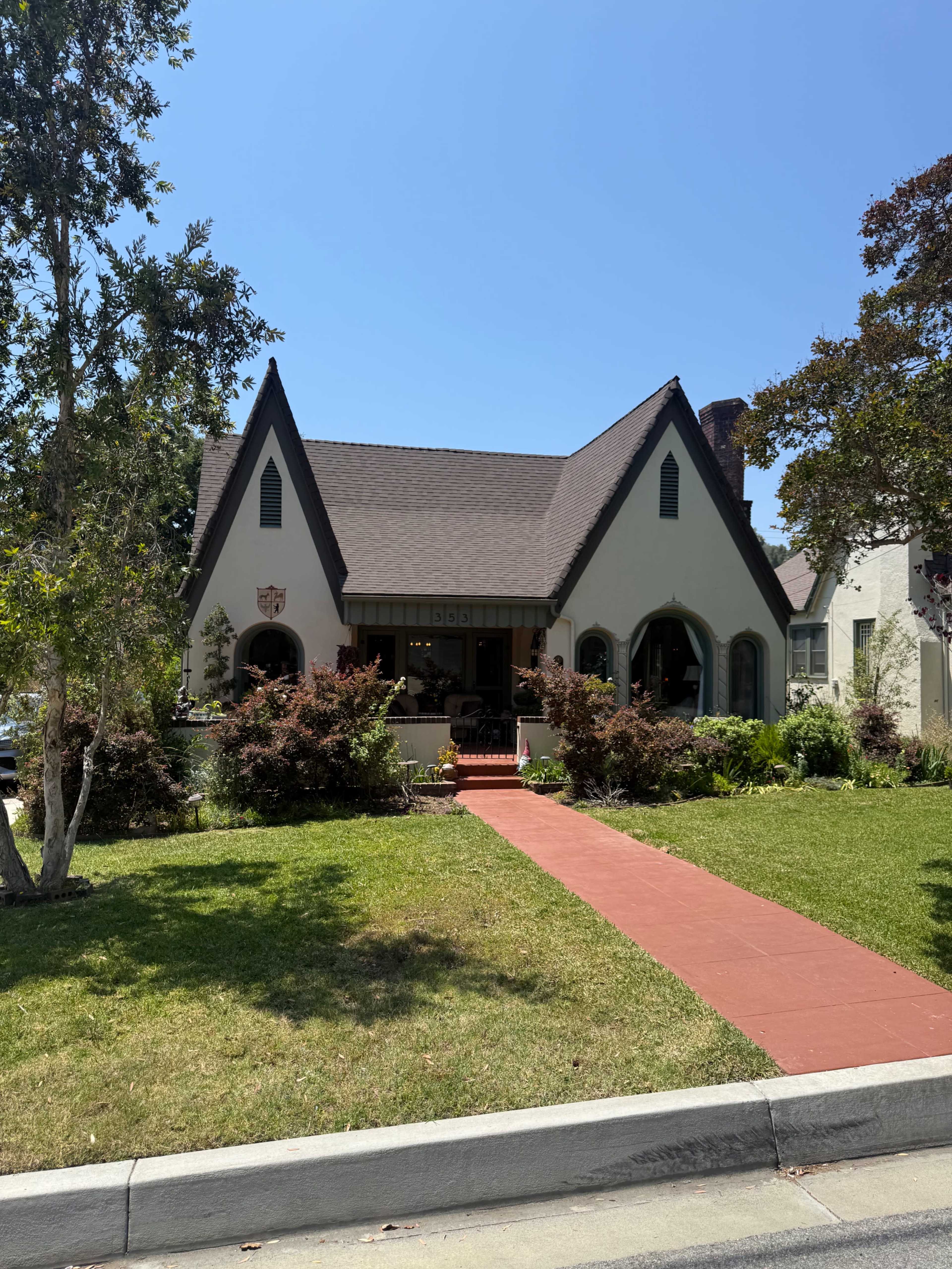 A quaint house with a peaked roof and a red brick walkway is surrounded by well-maintained shrubs and trees.