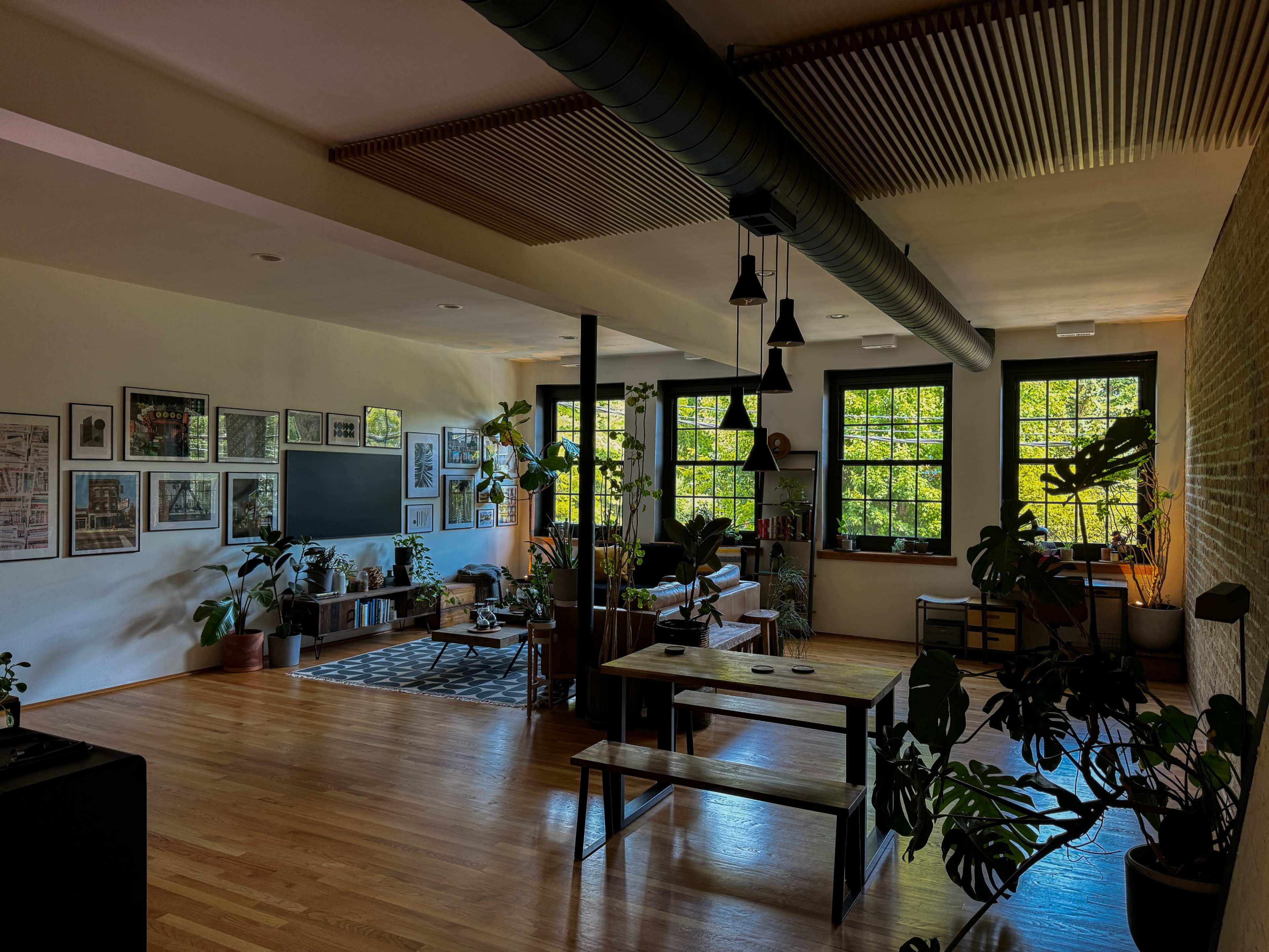 The image depicts a spacious, well-lit living area with large windows, wooden flooring, multiple plants, and a combination of seating and dining furniture.