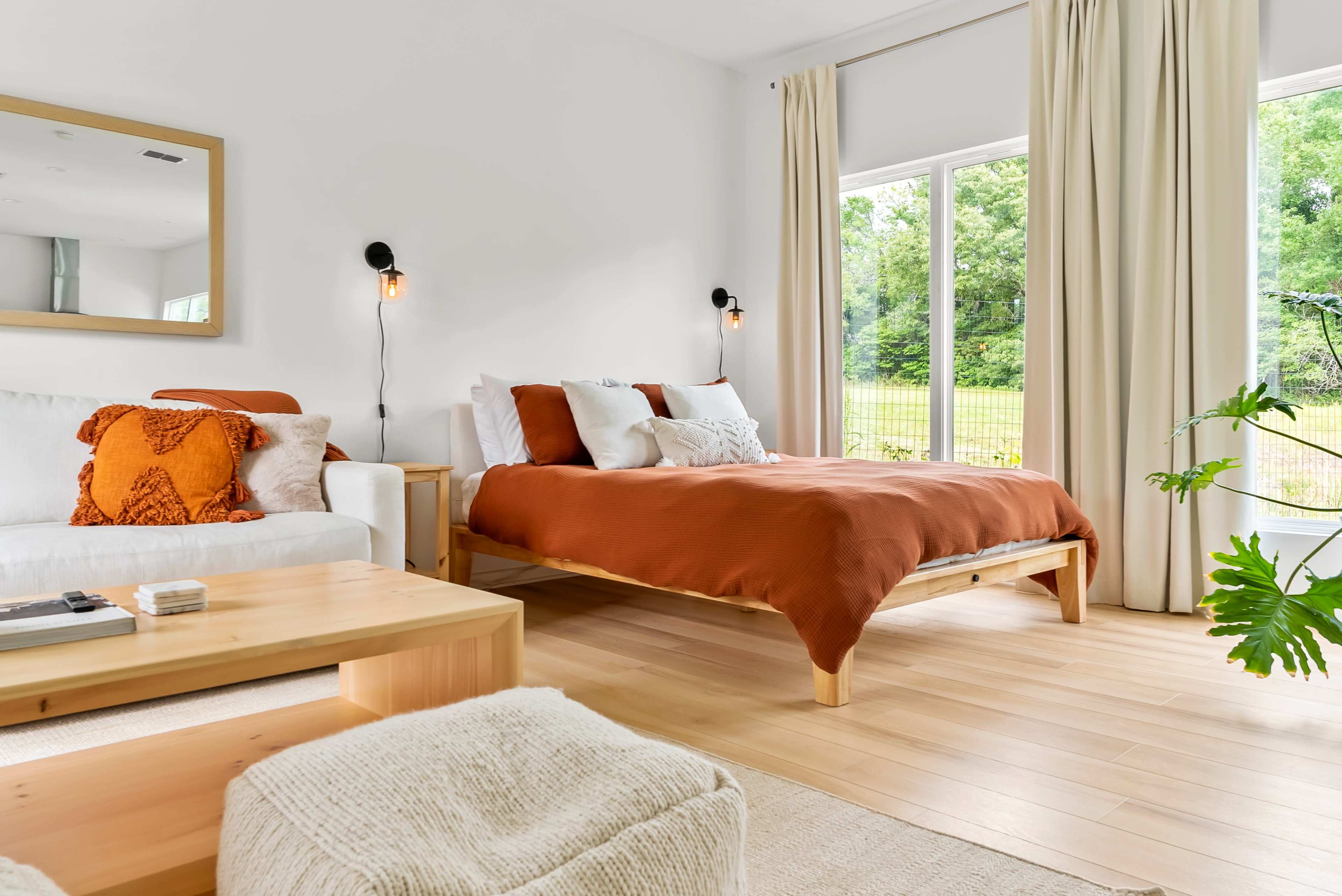 The bright bedroom features a wooden bed with orange bedding, a white sofa, and large windows overlooking a green outdoor area.