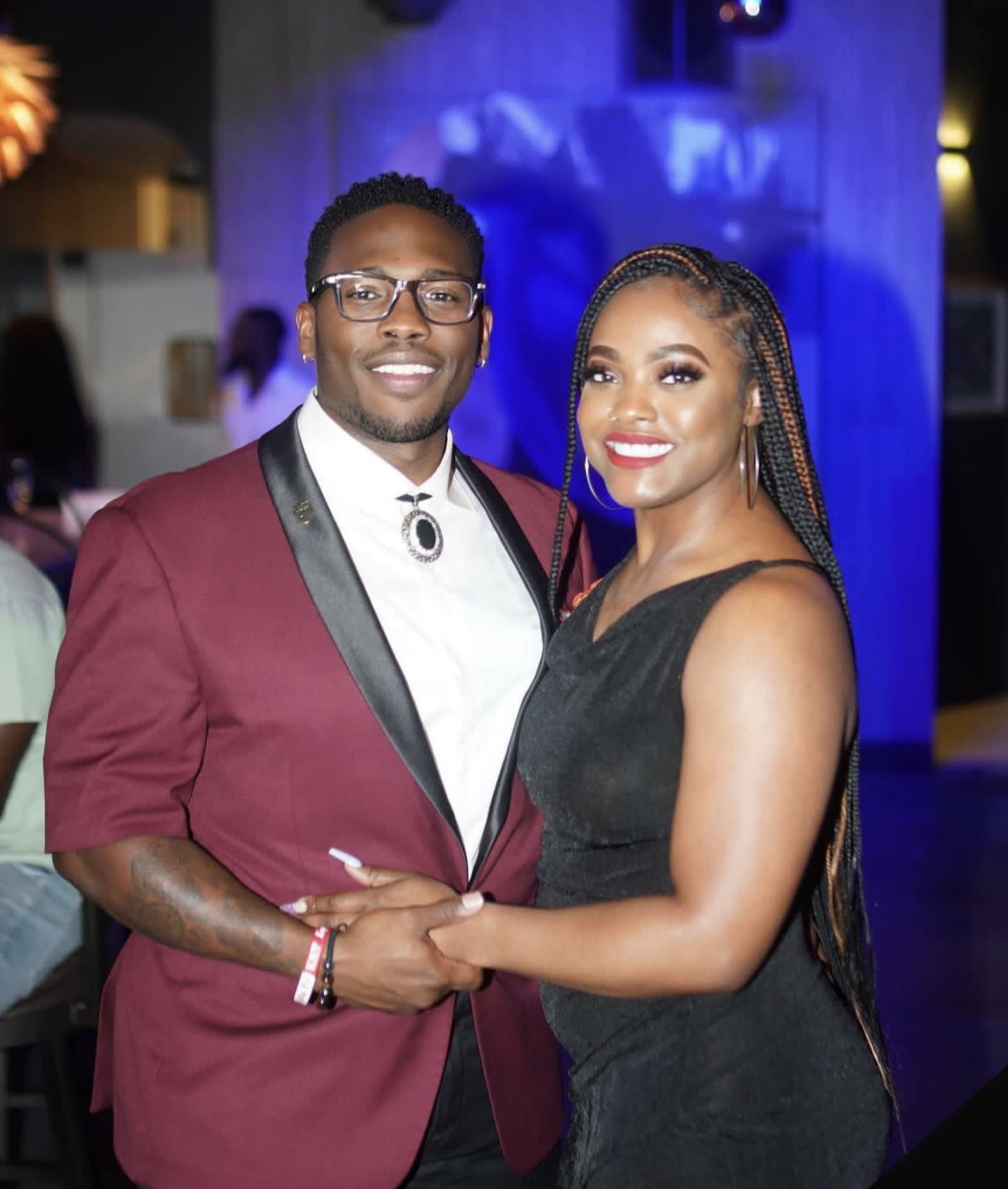 A man and a woman stand closely together in a dimly lit venue, both smiling at the camera.