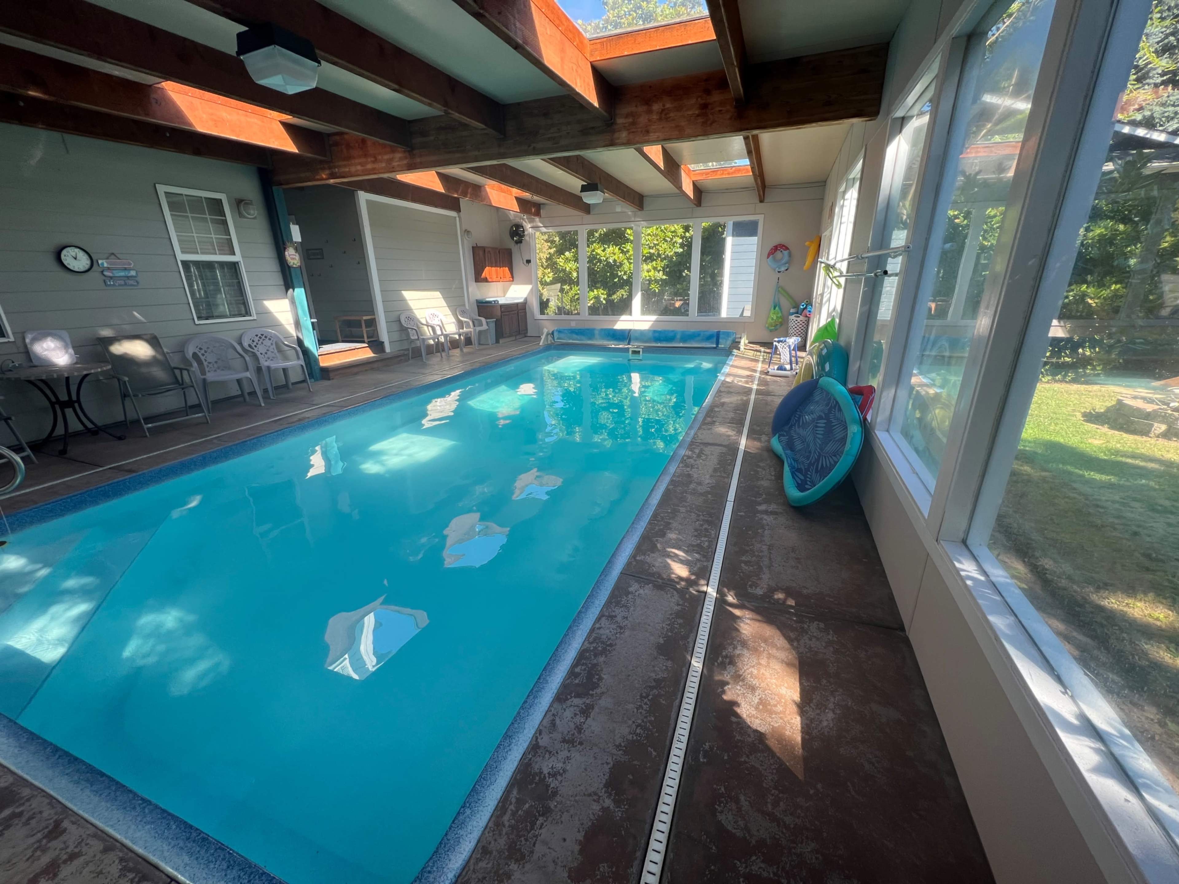 An indoor swimming pool surrounded by large windows and wooden beams, with a seating area beside it.