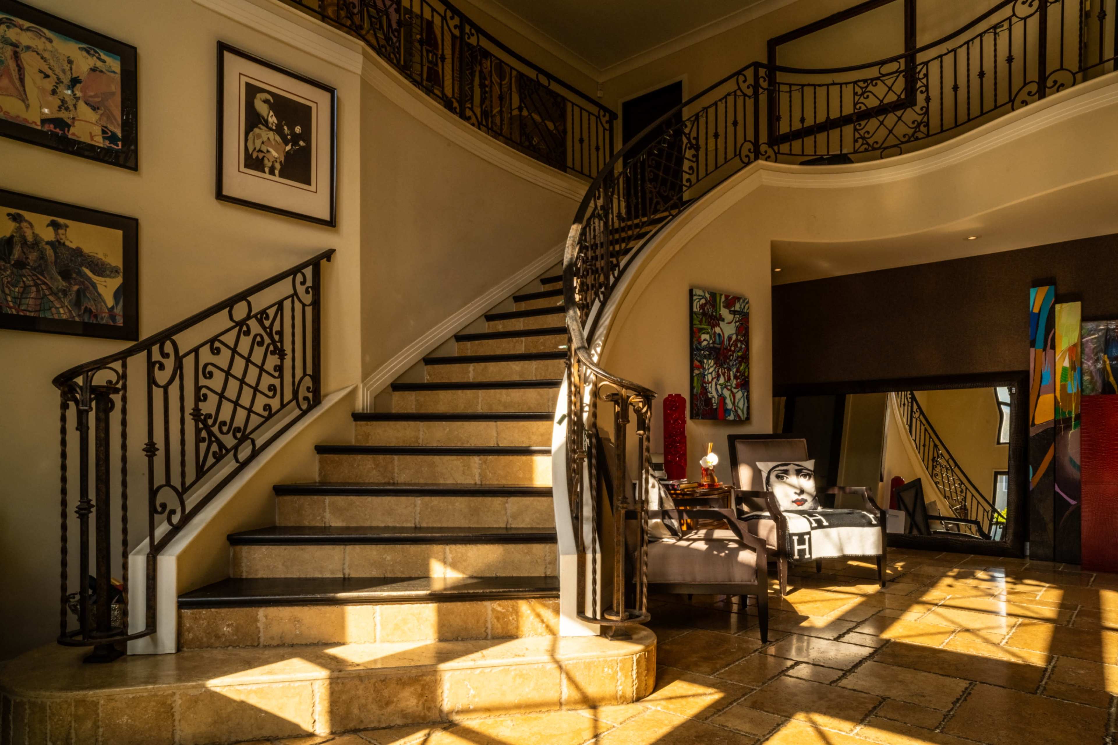 The image features a sunlit indoor staircase with wrought iron railings, leading to a second floor, surrounded by artworks on the walls and modern furniture in the foreground.