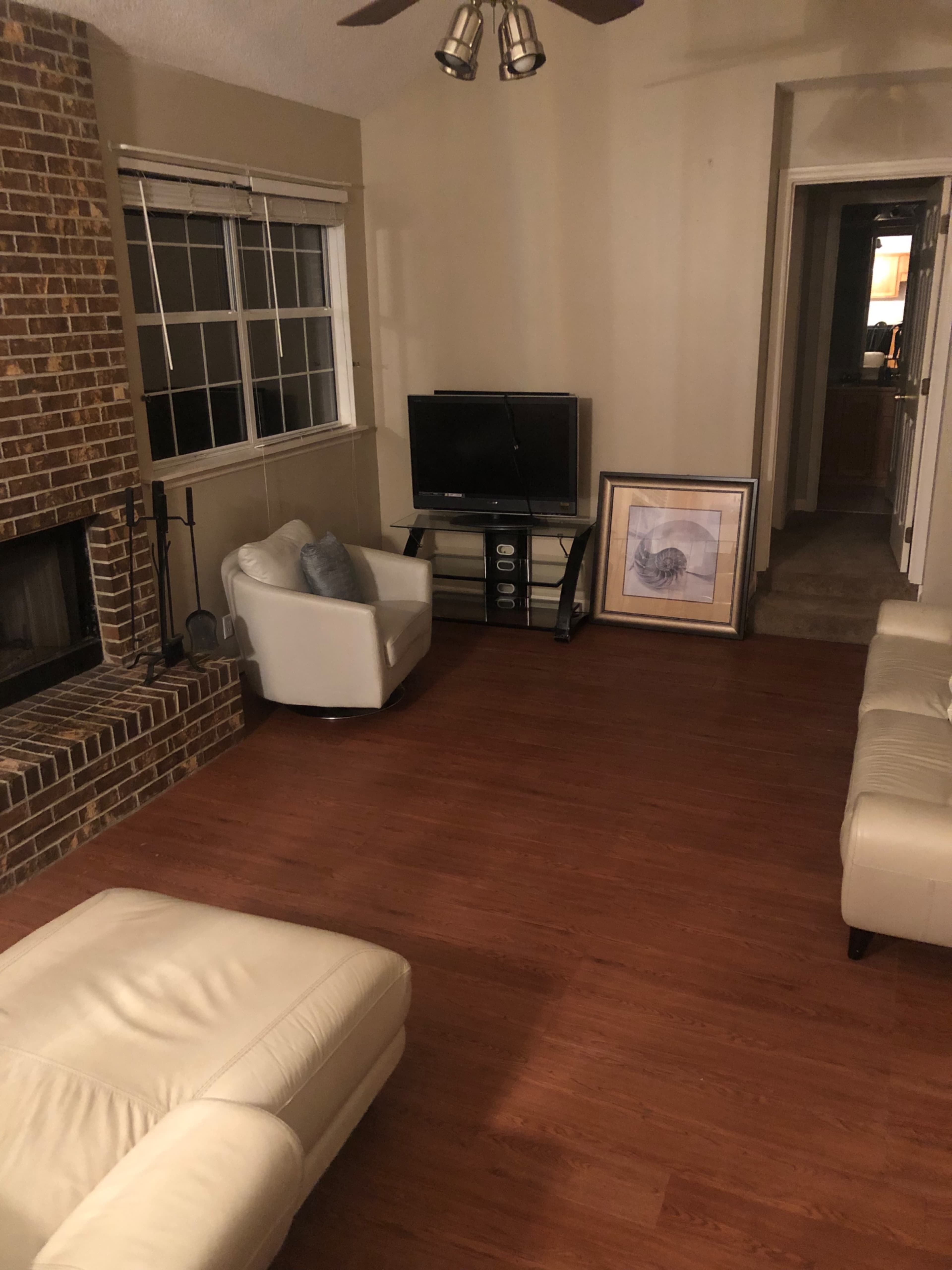 The living room features a brick fireplace, a television on a stand, two white chairs, and a framed picture leaning against the wall.