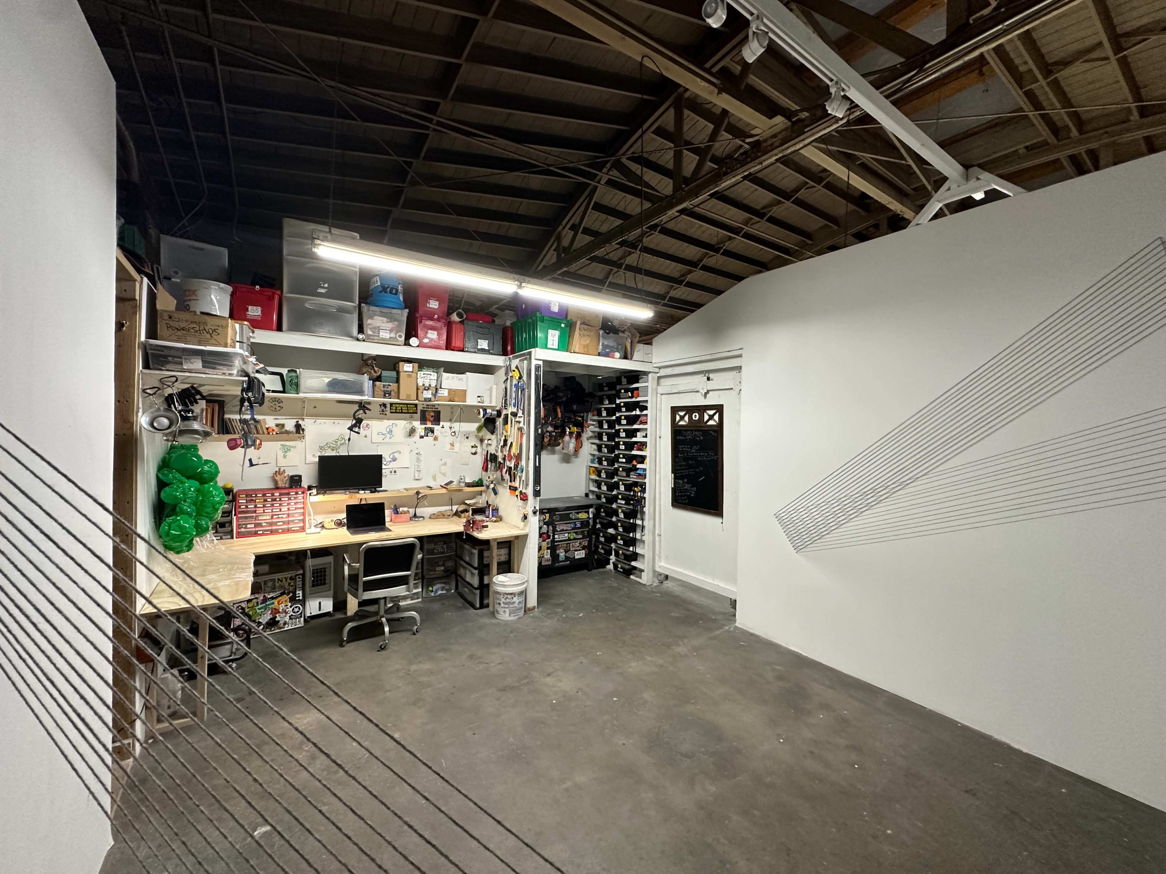 The image shows a workspace with a desk, computer, and various tools on shelves, situated in a garage-like environment with white walls and a line installation.