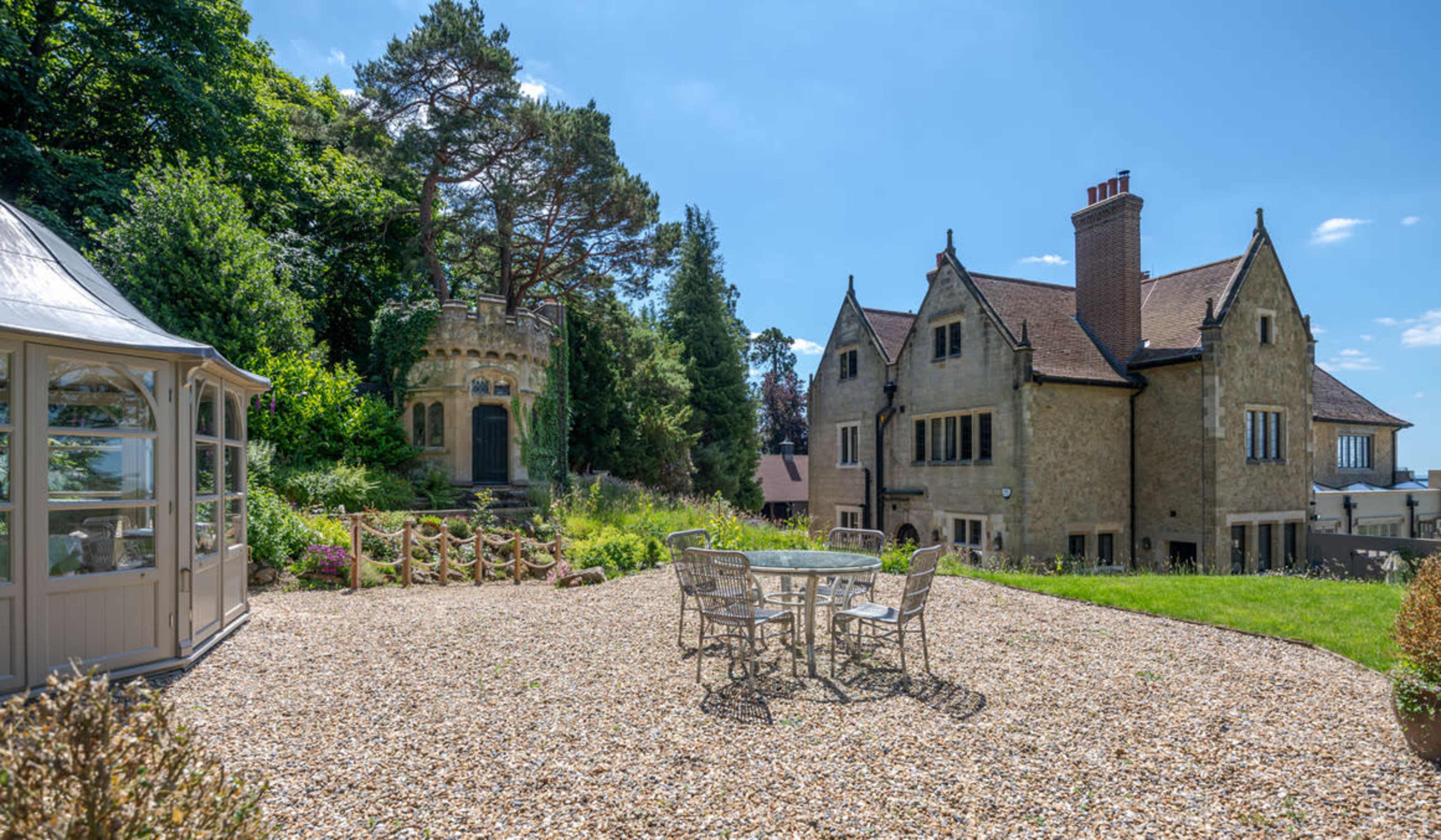 The image shows a spacious garden with a gravel area and a table surrounded by chairs, adjacent to a large stone house and a tower structure.