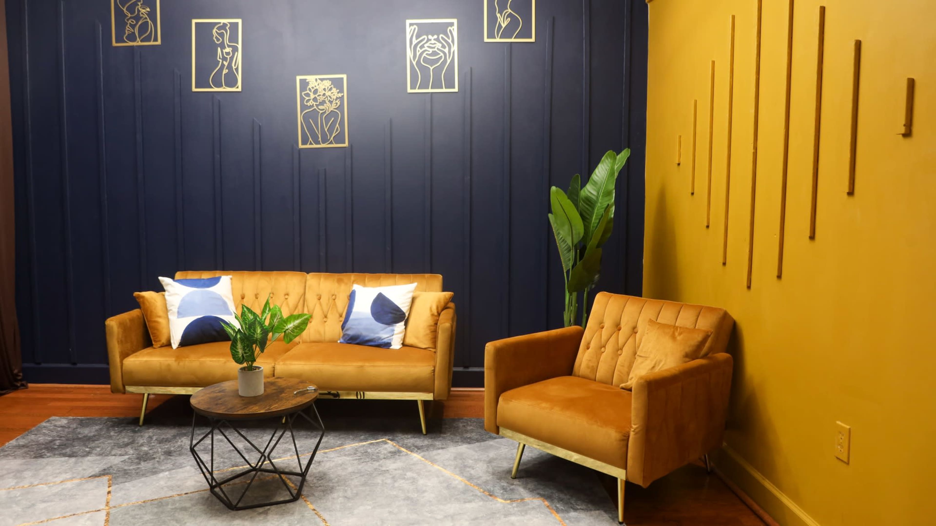 A modern sitting area with a mustard-yellow wall, navy-blue accents, a brown velvet sofa and armchair, a round coffee table, and decorative wall art.