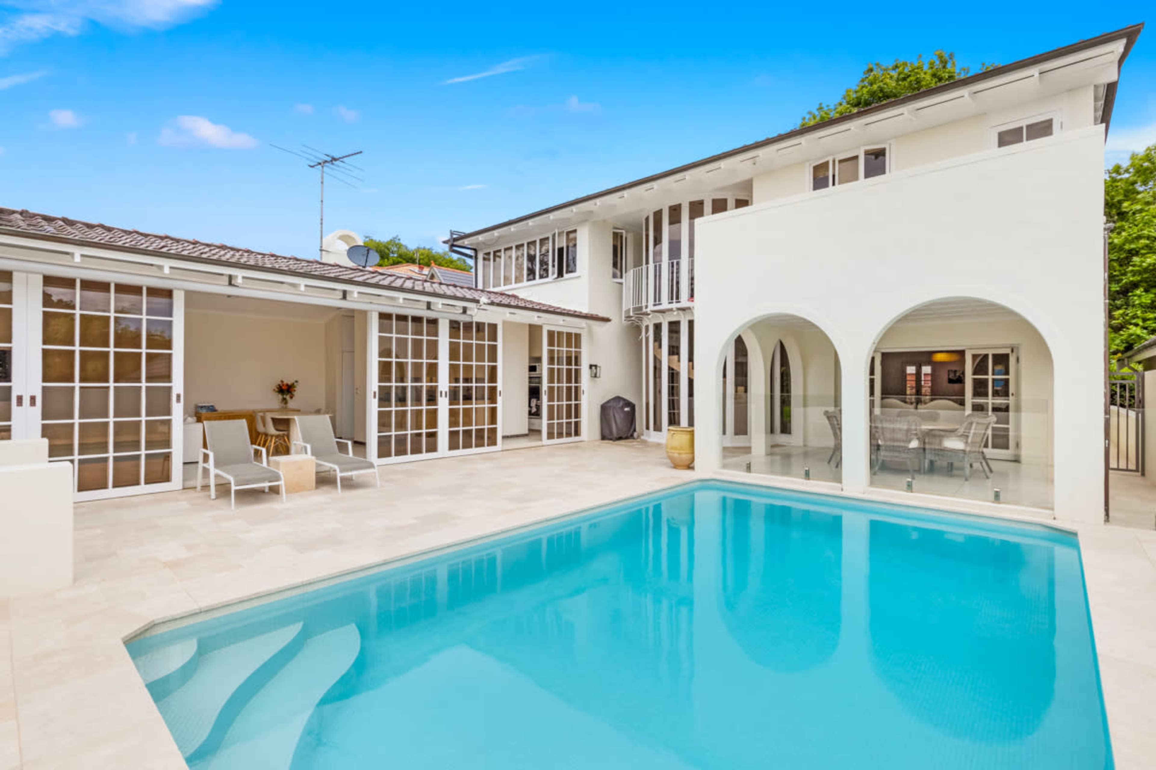 The image features a modern house with a swimming pool, surrounded by a tiled patio and large glass doors.