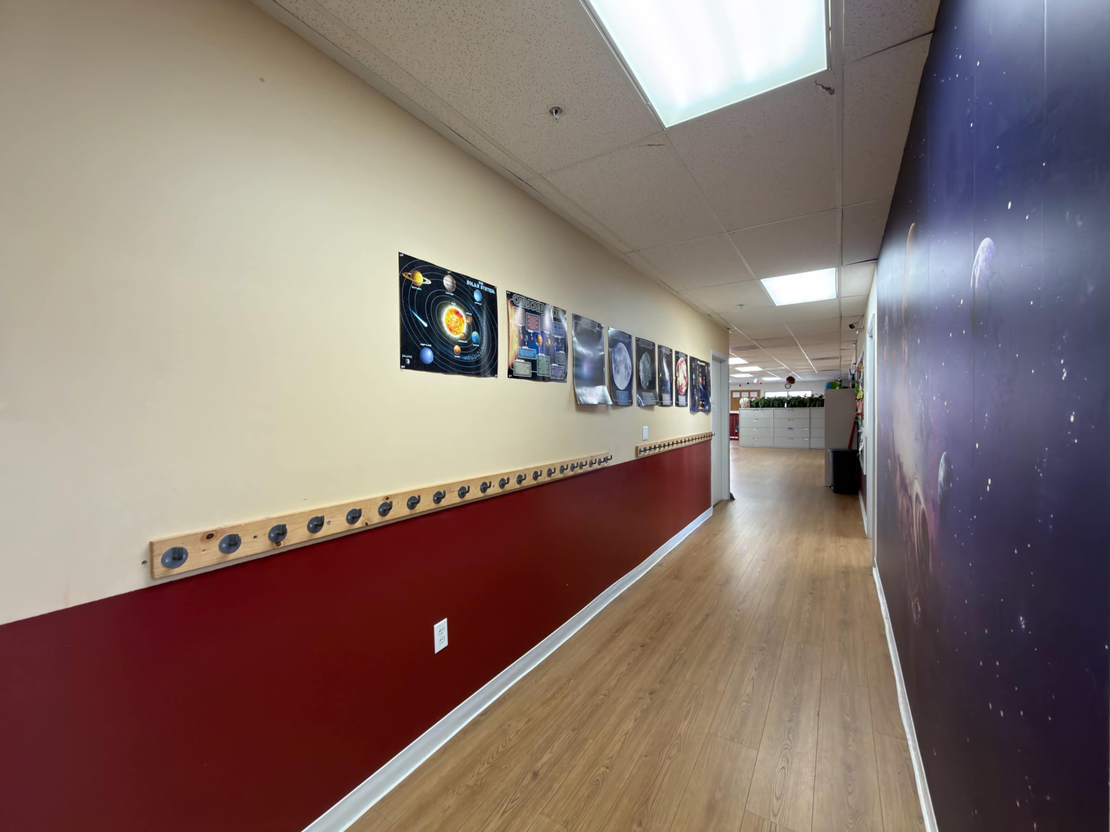 The image shows a hallway with a tan and maroon wall, featuring framed pictures on one side and a section of the ceiling illuminated by fluorescent lights.