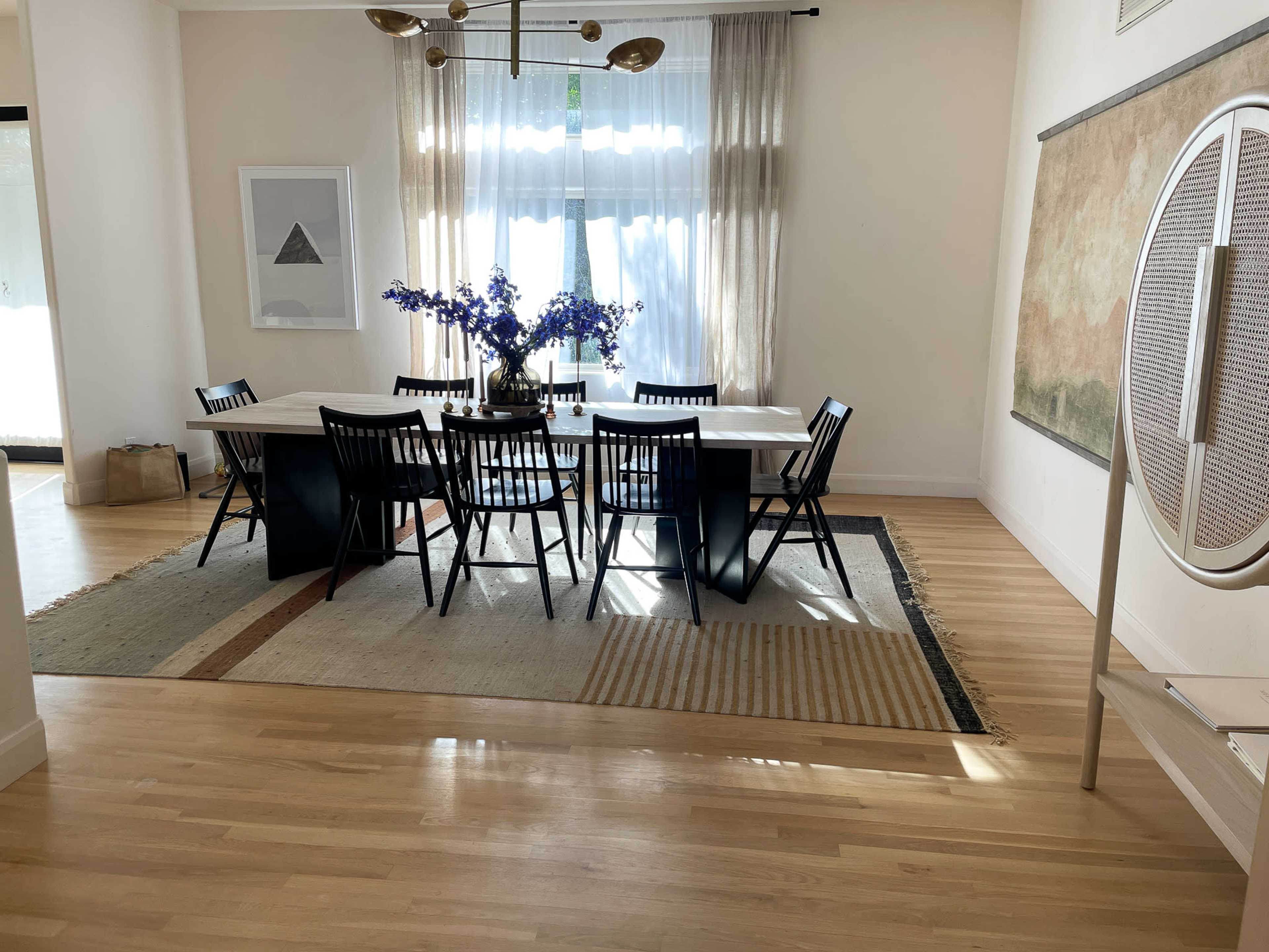 A dining room features a long table surrounded by black chairs, with a vase of purple flowers at the center, set on a patterned rug under natural light from large windows.