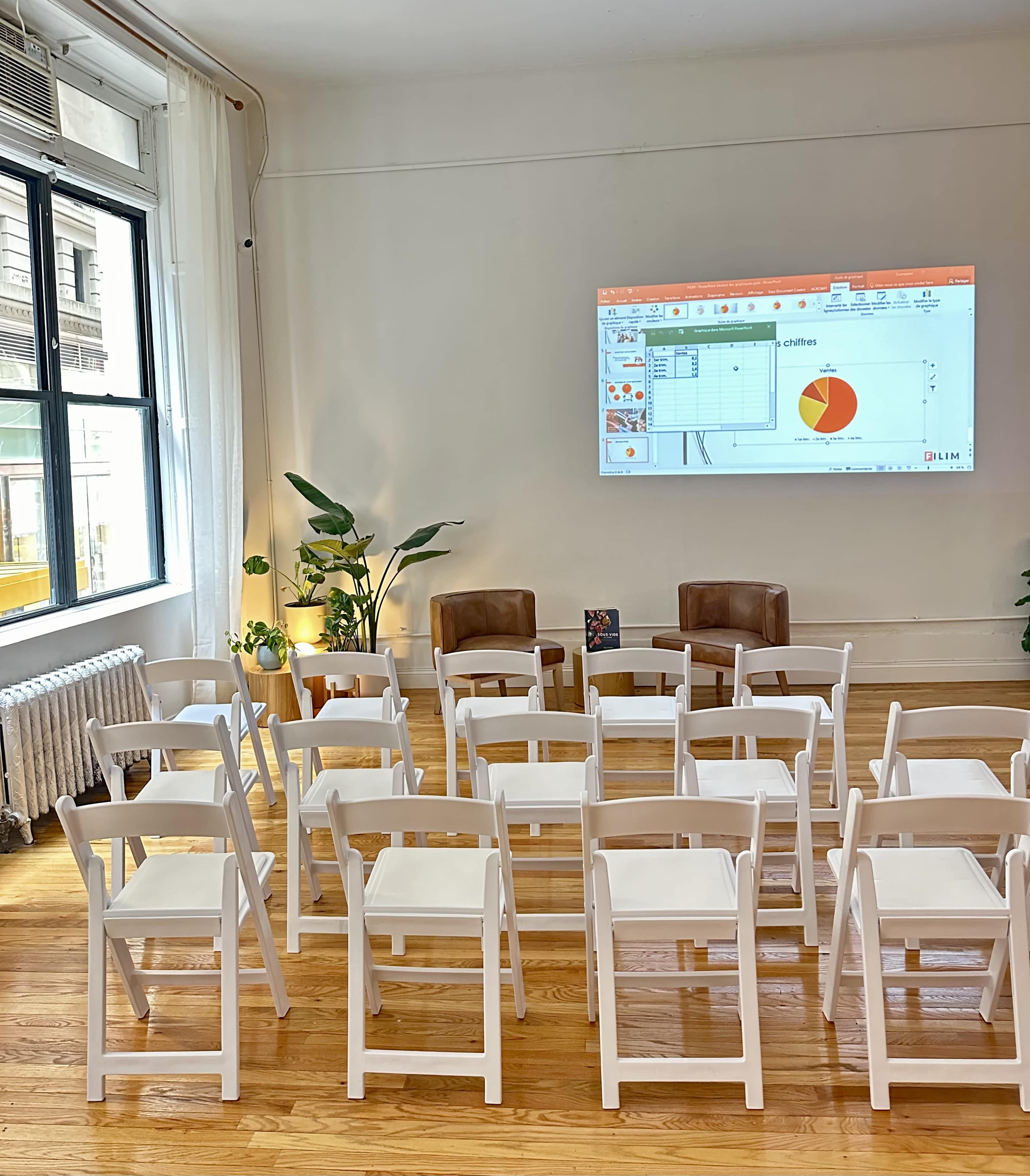 A room with white chairs arranged in rows facing a projector screen displaying a chart, with two brown chairs and a potted plant in the background.