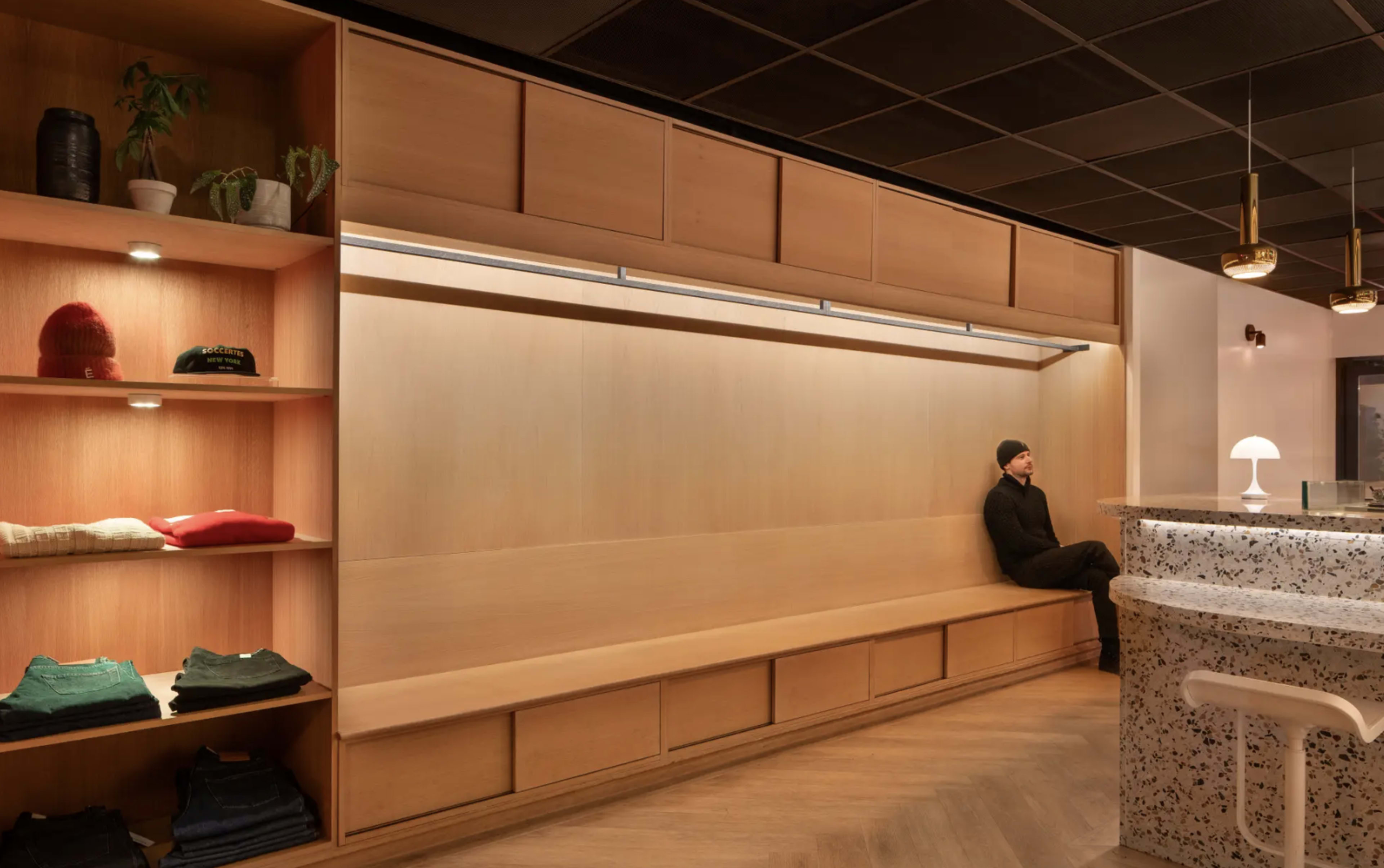 The image shows a minimalist interior space with a wooden bench along one wall, a display shelf with neatly folded clothing, and a person sitting on the bench.