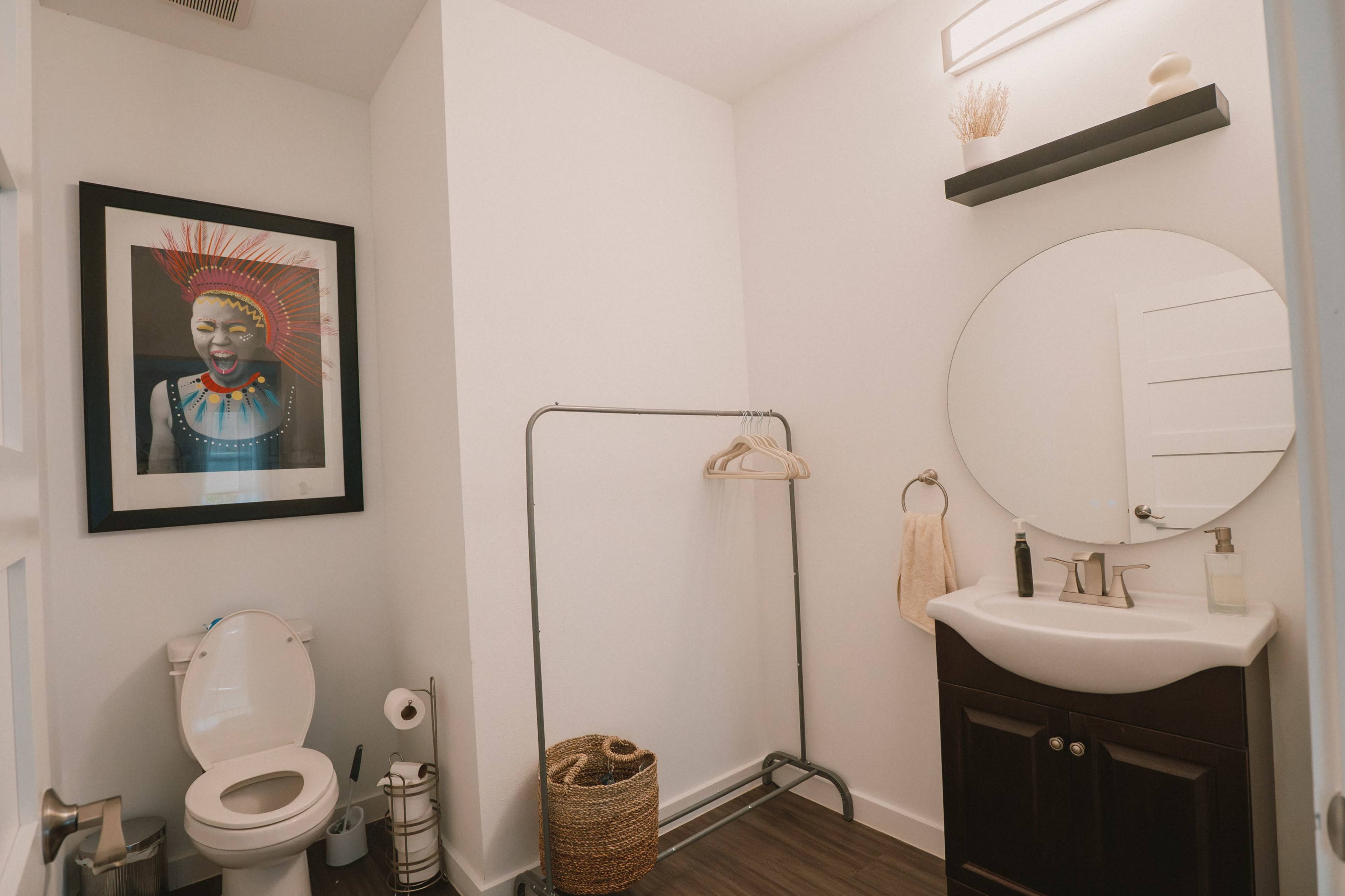 The image shows a small bathroom featuring a toilet, a sink with a mirror, a clothing rack, and a framed artwork on the wall.