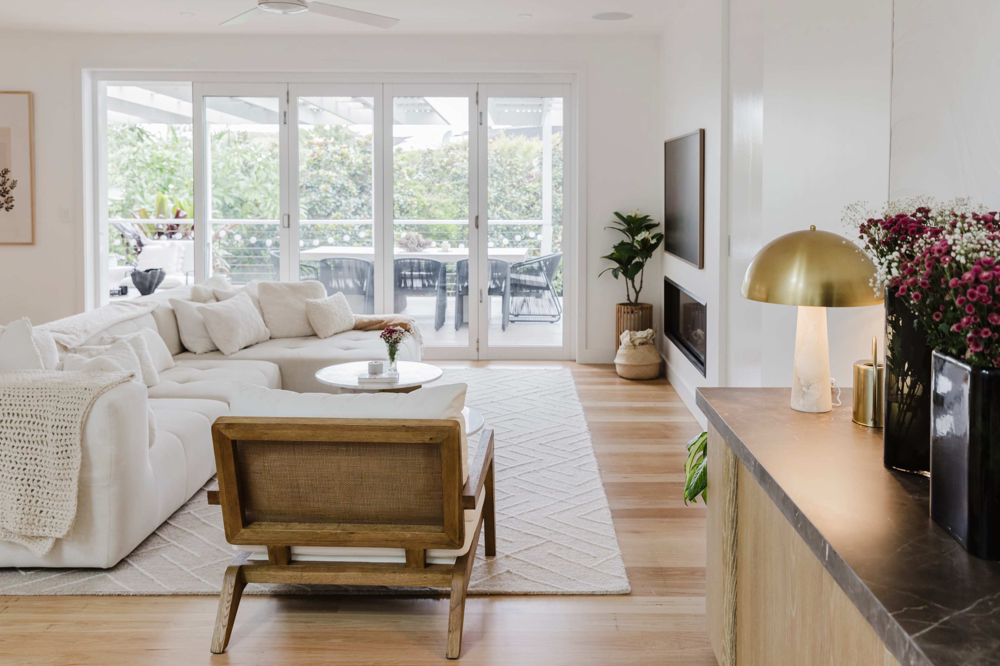 The image shows a modern living room with a large sectional sofa, a coffee table, and a comfortable chair, featuring large windows that provide a view of a green outdoor space.