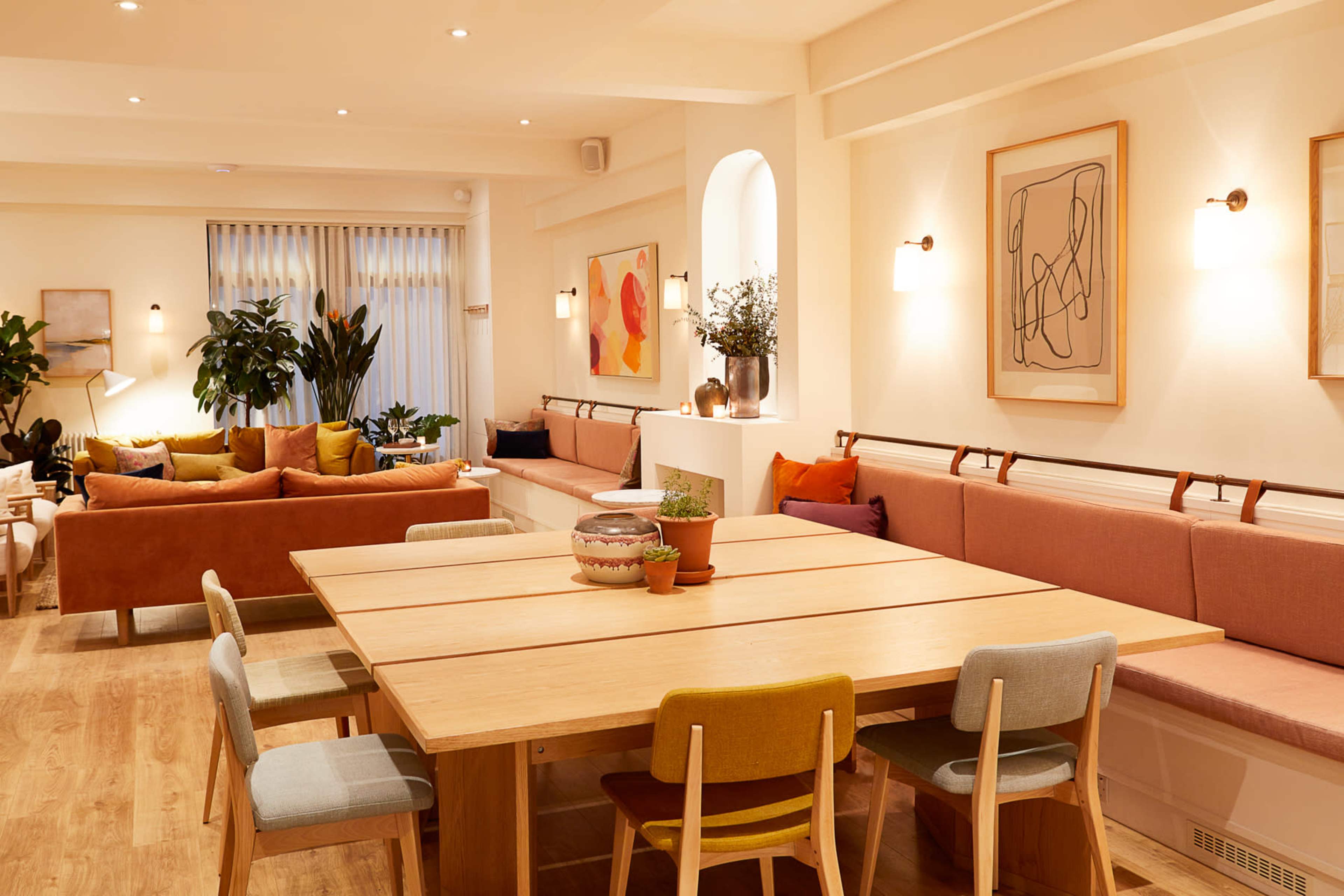 A spacious, modern interior with a large wooden dining table surrounded by colorful chairs, complemented by lounge seating and plants along the walls.
