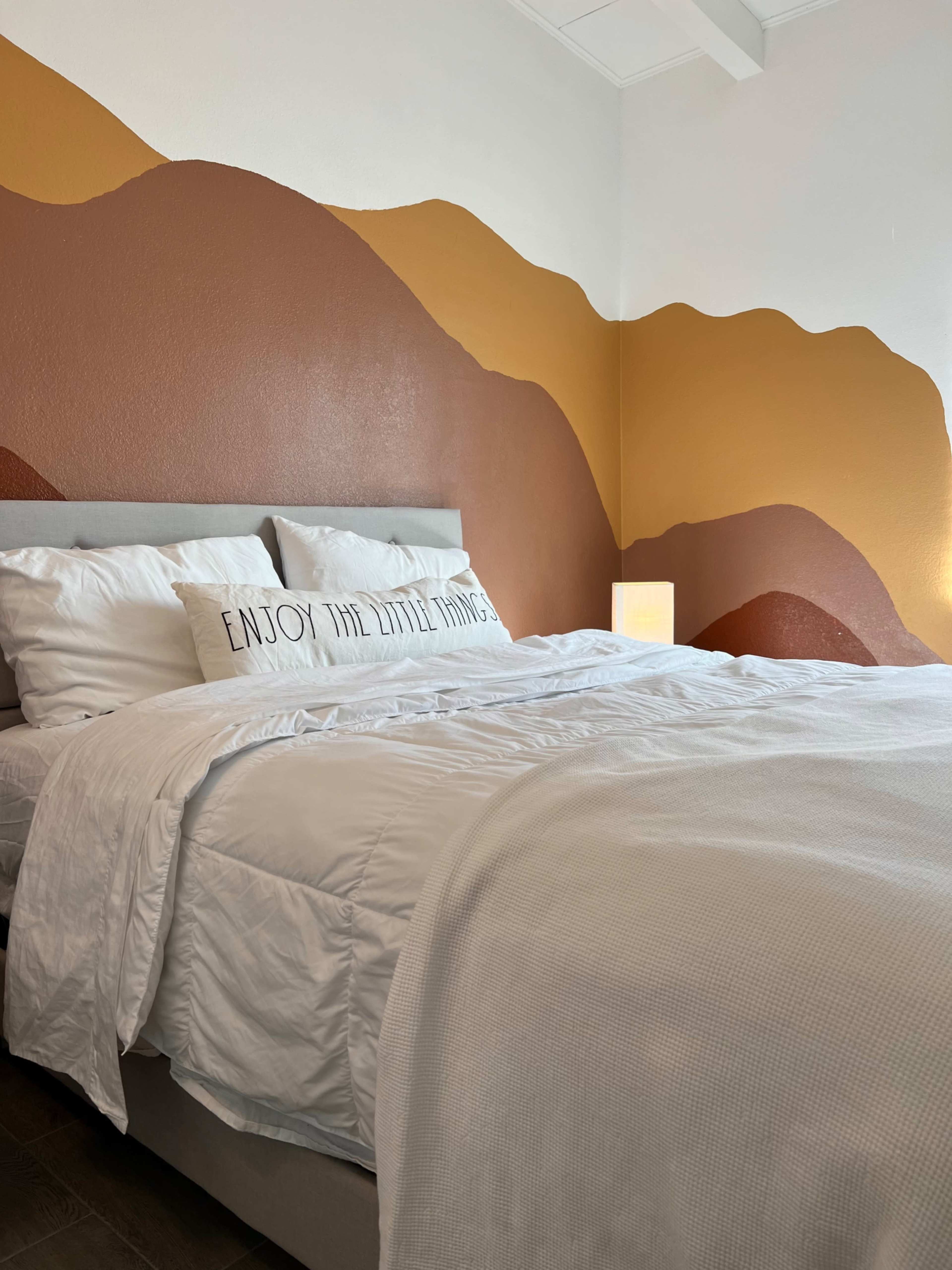 The image shows a neatly made bed with white bedding and a decorative pillow in a room featuring a brown and gold abstract mural on one wall.