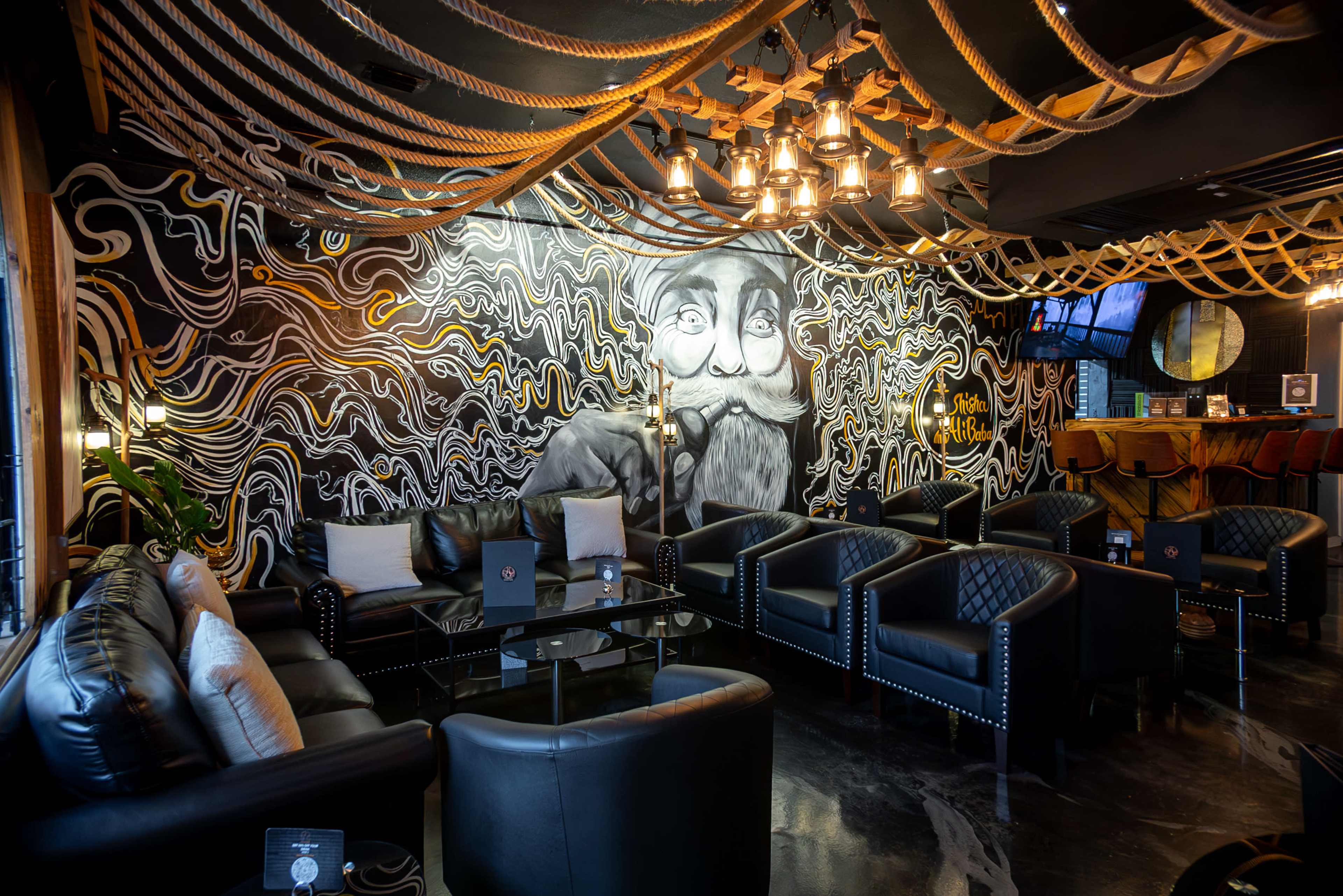 The image shows a stylish lounge with black leather seating, a mural of a man with a mustache on the wall, and decorative ropes hanging from the ceiling.