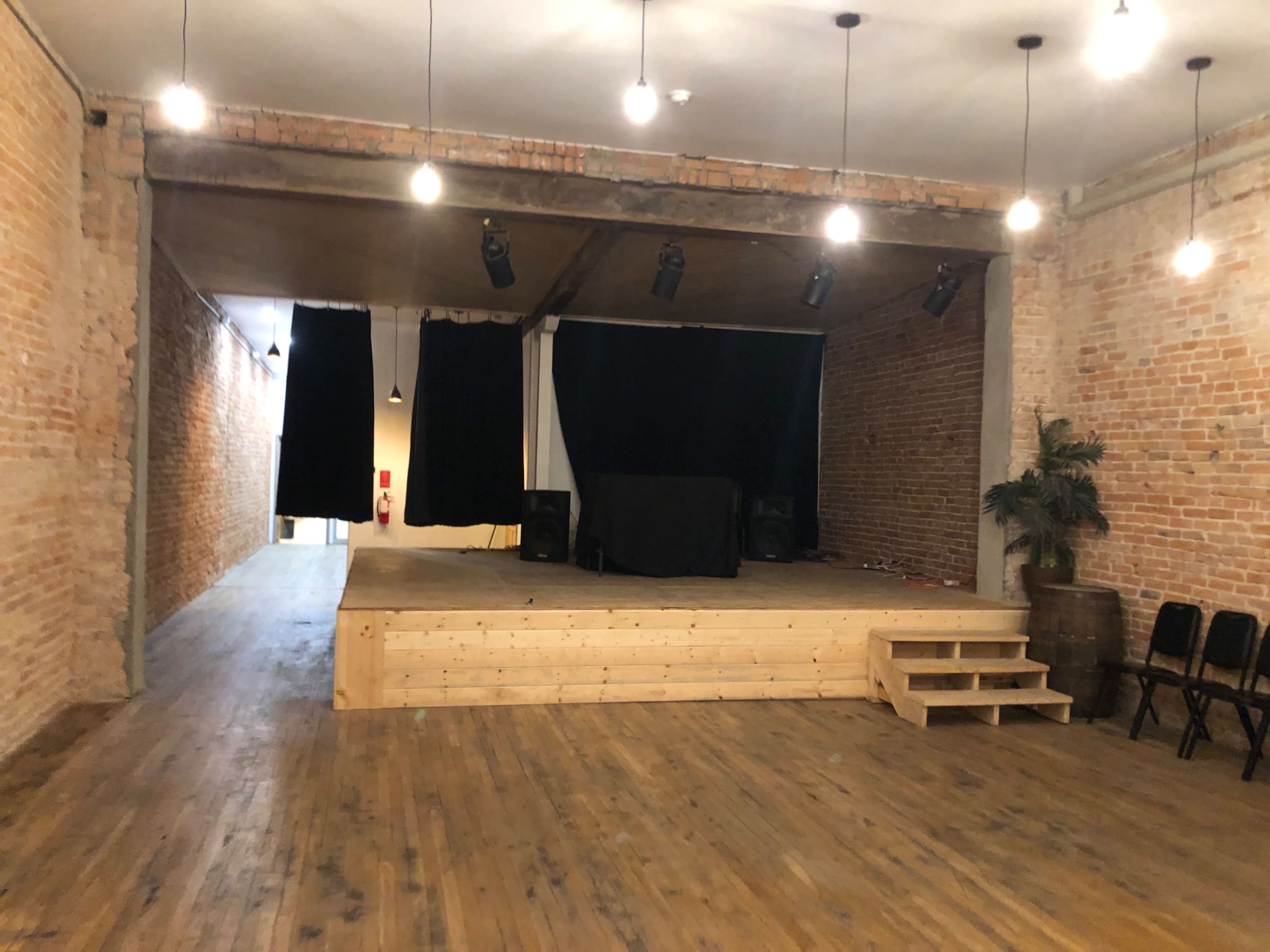 An empty performance space with a wooden stage, brick walls, and overhead lighting.