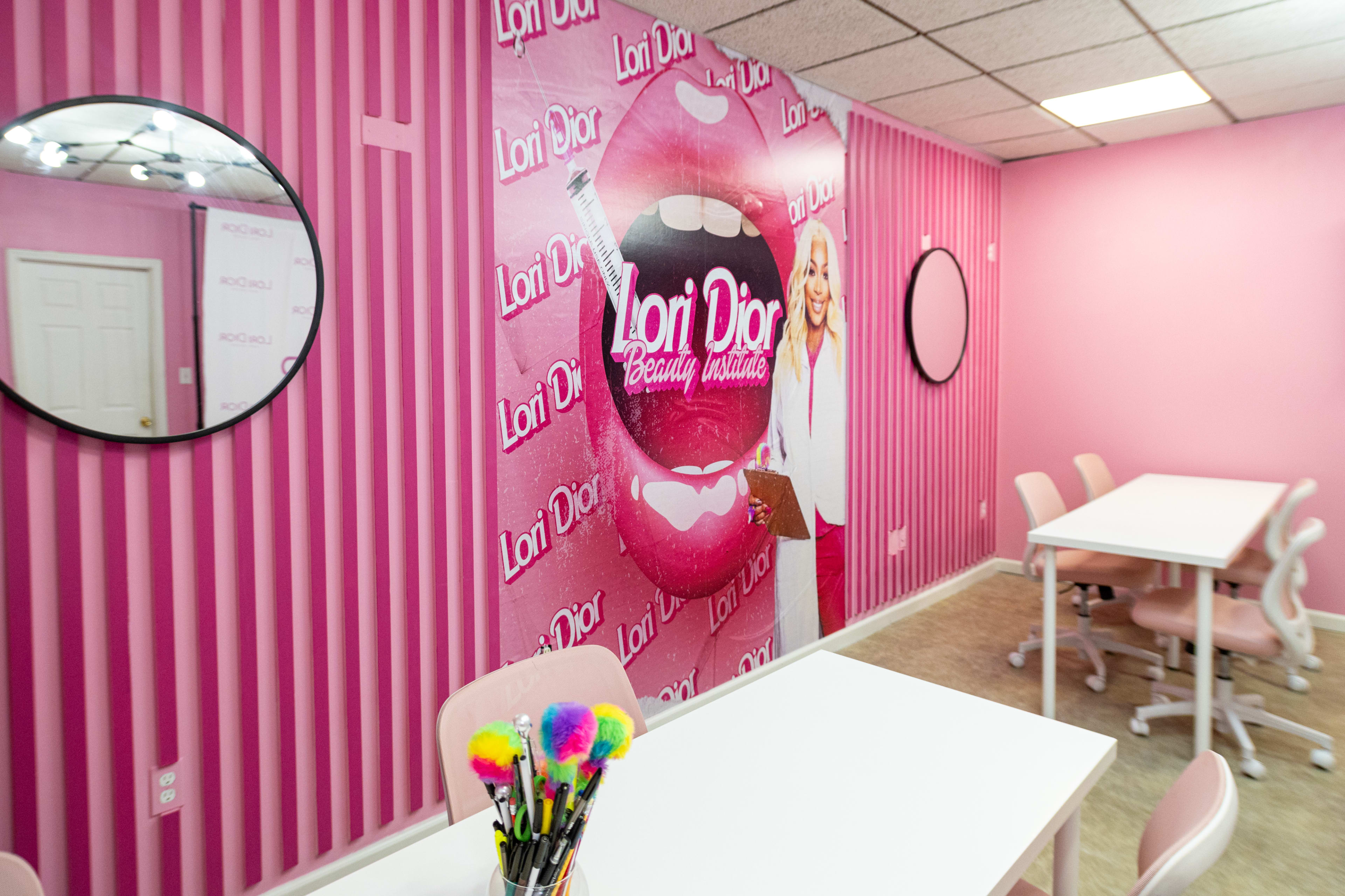 A brightly colored salon interior featuring pink striped walls with branding, two round mirrors, and white tables surrounded by chairs.