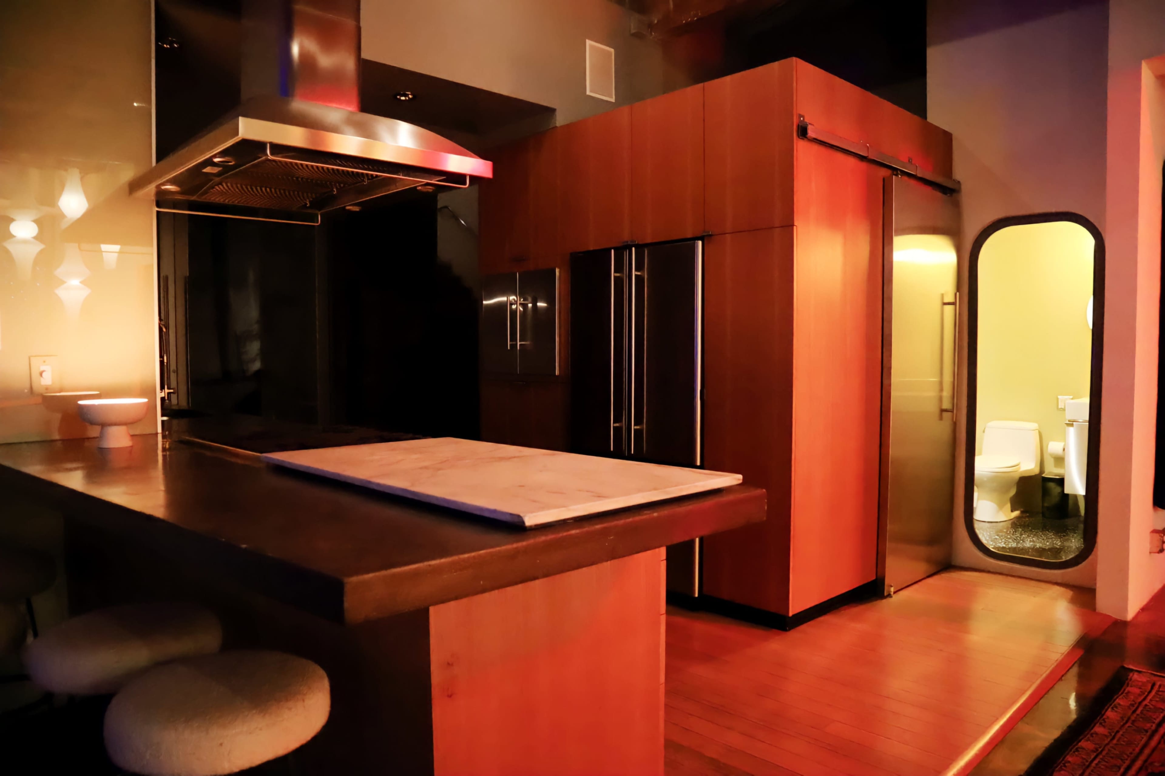 The image shows a modern kitchen with wooden cabinetry, a large island countertop, and an open doorway leading to a bathroom.